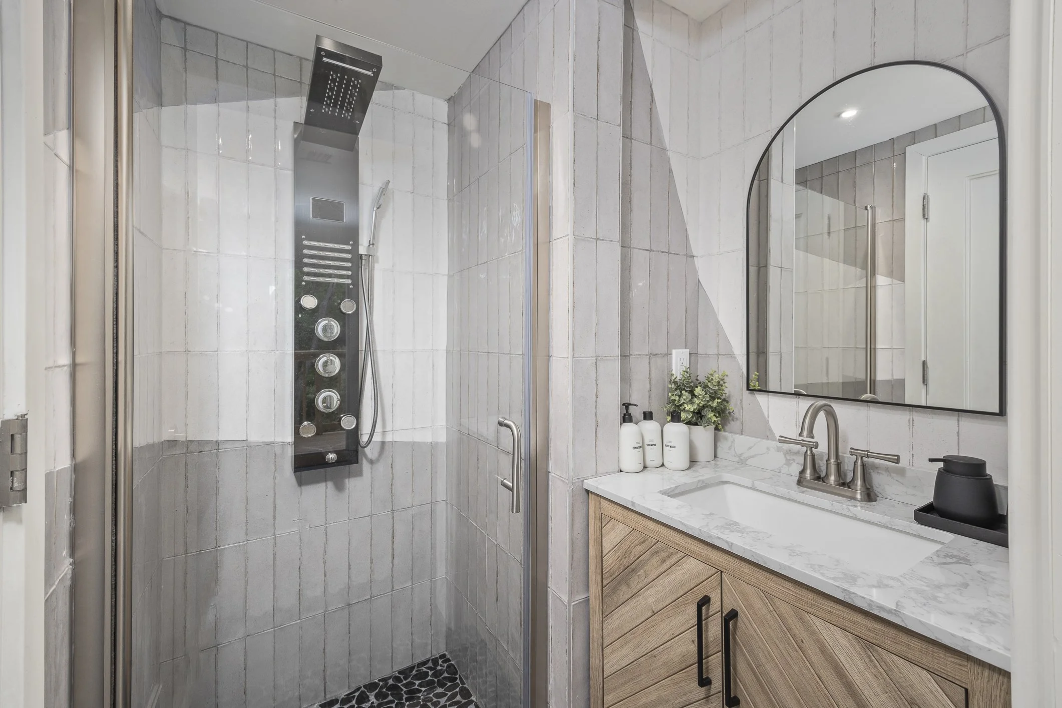 Modern bathroom with a walk-in shower featuring a digital shower panel, a marble countertop vanity with a rectangular sink, a large mirror, and decorative soap dispensers and a plant.