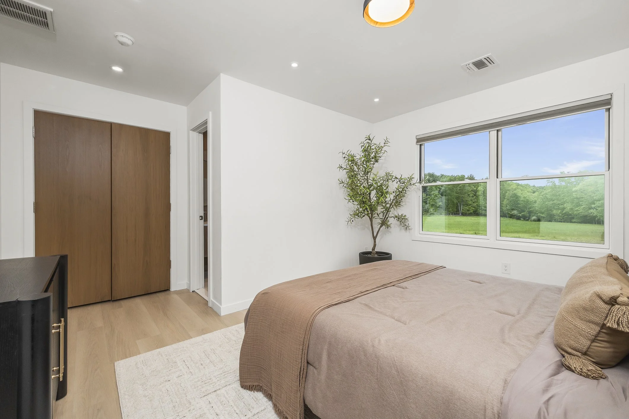 A bedroom with a bed covered in beige and brown bedding, a large window showing a green landscape outside, a potted plant near the window, a closed wooden closet, and a white wall.