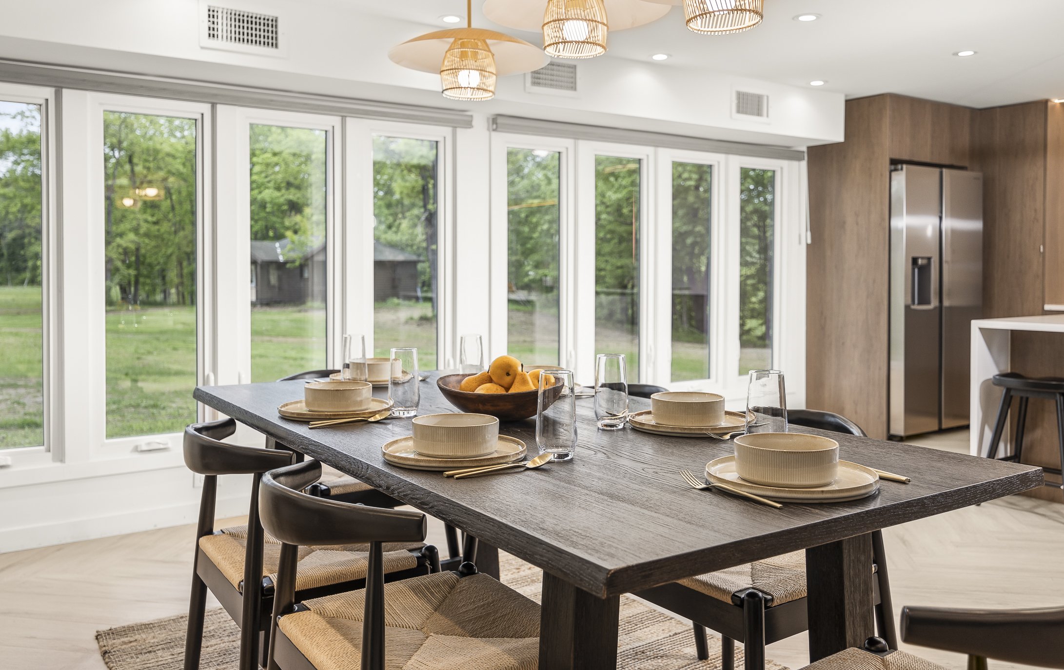 A modern dining room with a wooden table set for six, featuring beige bowls, plates, and gold utensils, with a bowl of peaches in the center. Large windows showcase an outdoor green landscape, and hanging wicker pendant lights illuminate the space.