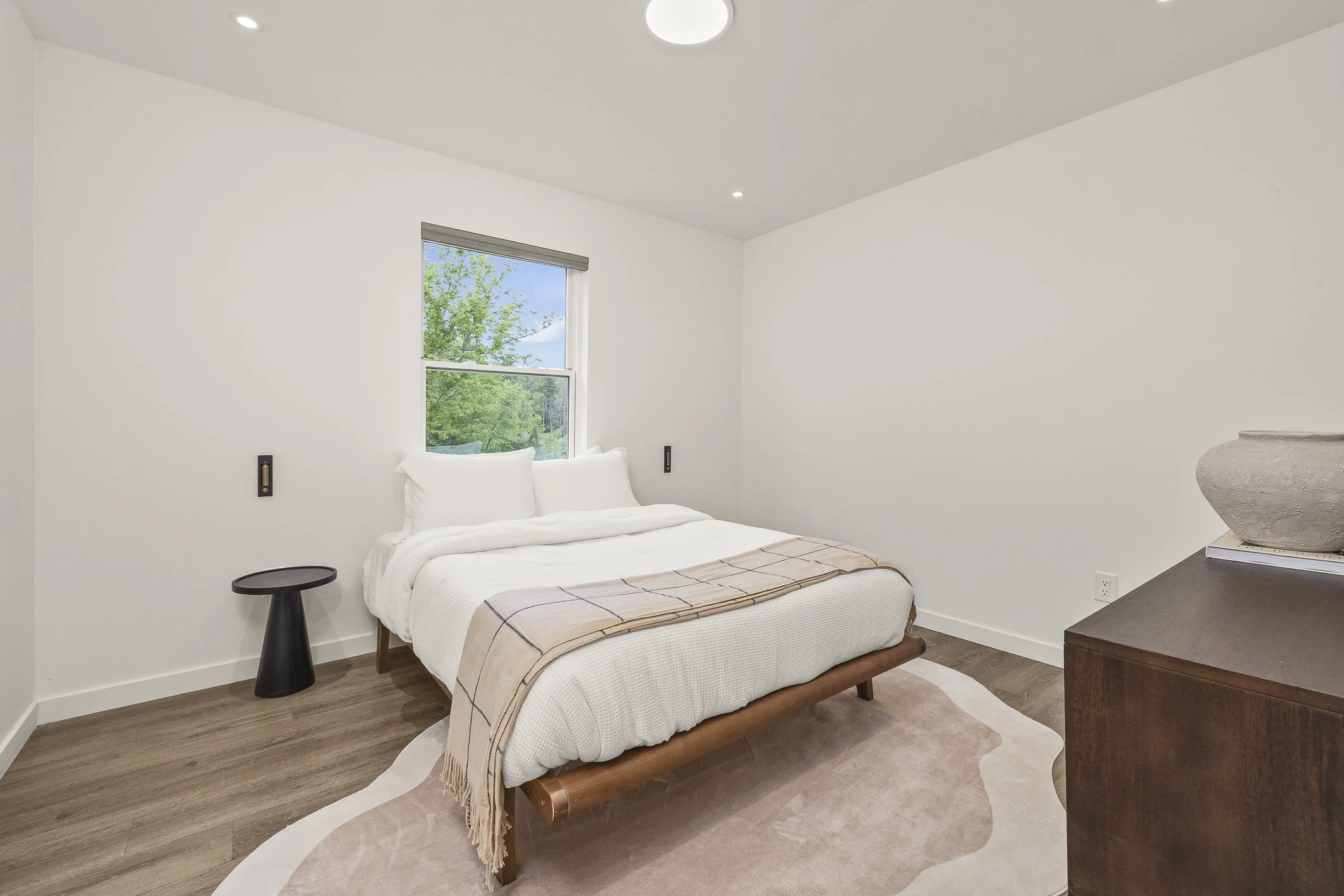 Minimalist bedroom with white walls, a bed with white bedding and pillows, a window showing green trees outside, a dark side table, and a wooden dresser with a decorative bowl on top.