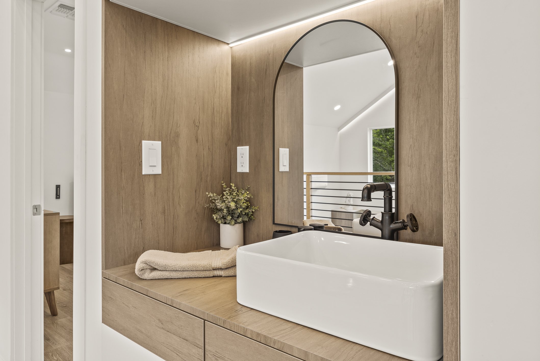Bathroom vanity area with a white rectangular vessel sink, black faucet, beige towels, green plant in a white pot, large arched mirror, wooden cabinet, and light-colored wall with integrated lighting.