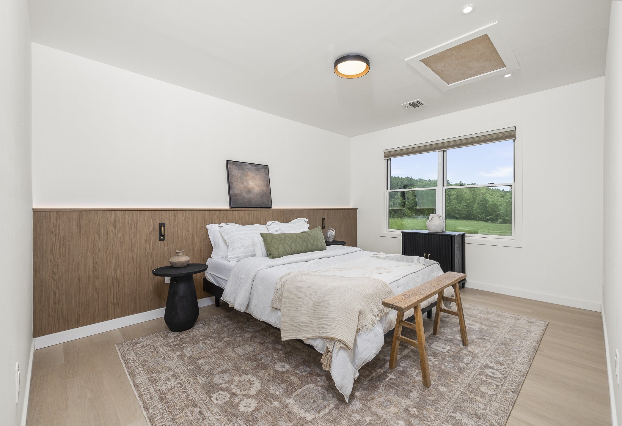 A minimalist bedroom with a bed against a wood-paneled headboard, a black side table, a bench at the foot of the bed, and a window showing a green outdoor landscape.