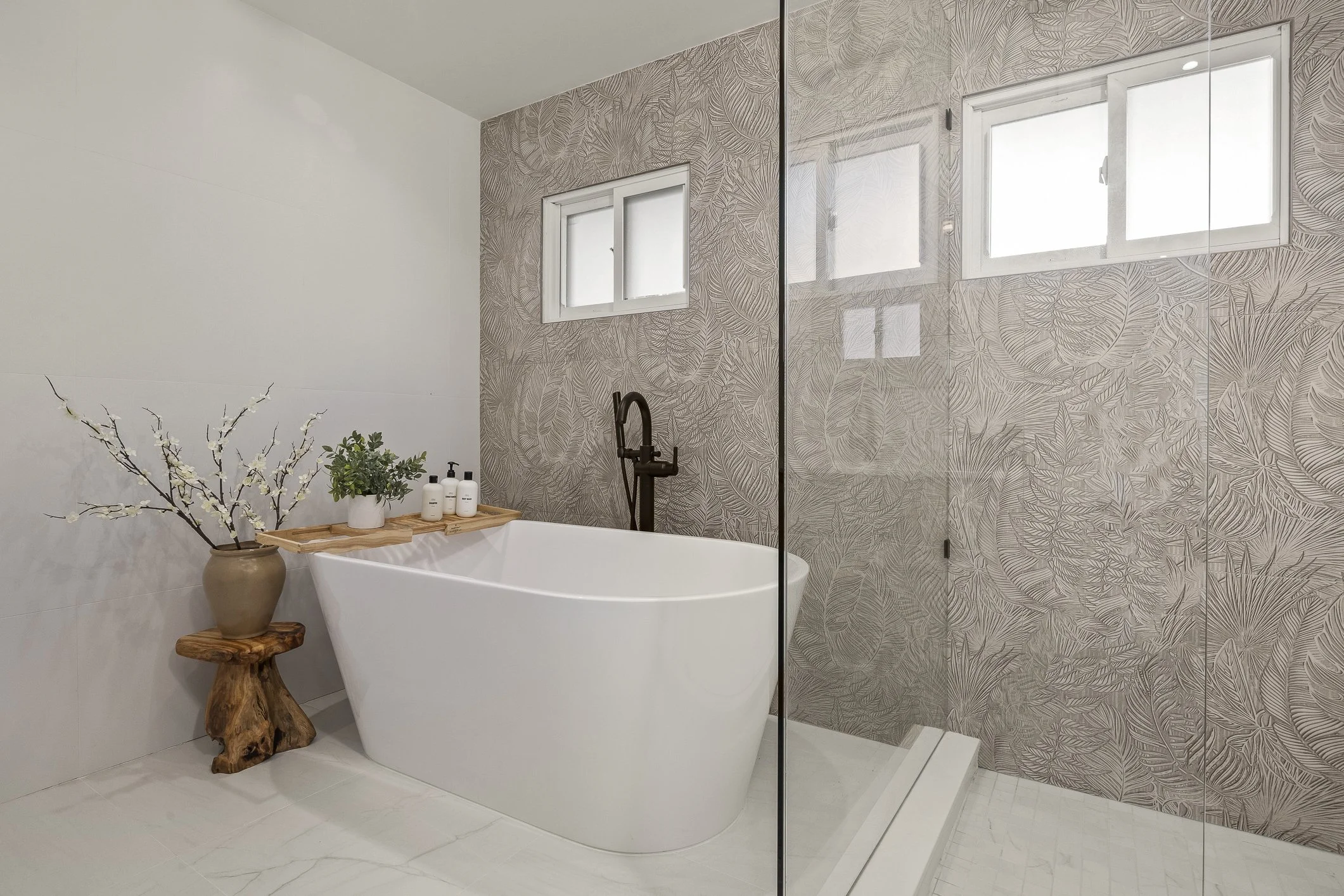 Modern bathroom with white bathtub, wooden side table with a potted plant and decorative branches, glass shower enclosure, and beige leaf-patterned wall tiles near two small windows.