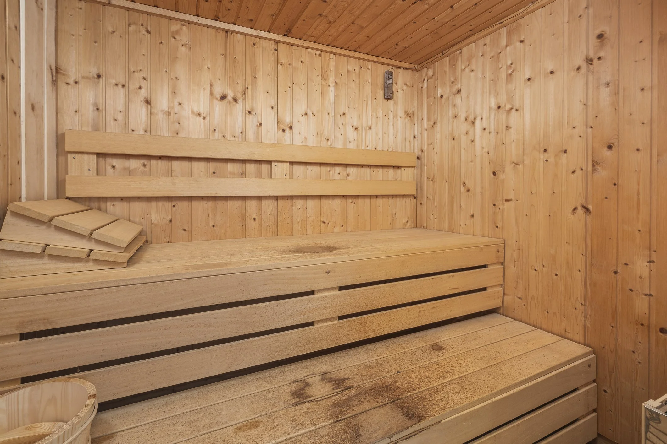 Interior of a wooden sauna with wooden benches, a small stack of wooden backrests, and a sauna bucket in the corner.