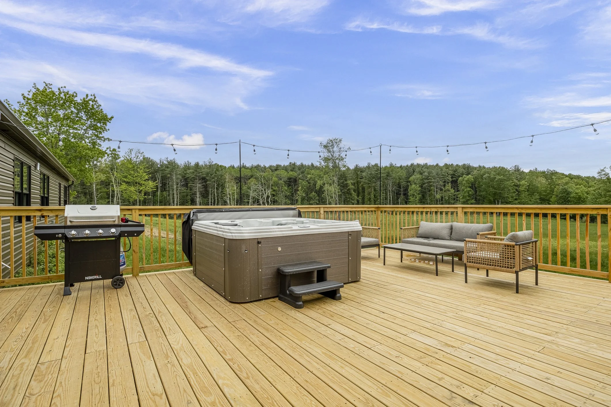 Wooden deck with outdoor furniture, hot tub, and grill, overlooking trees and a blue sky with some clouds.