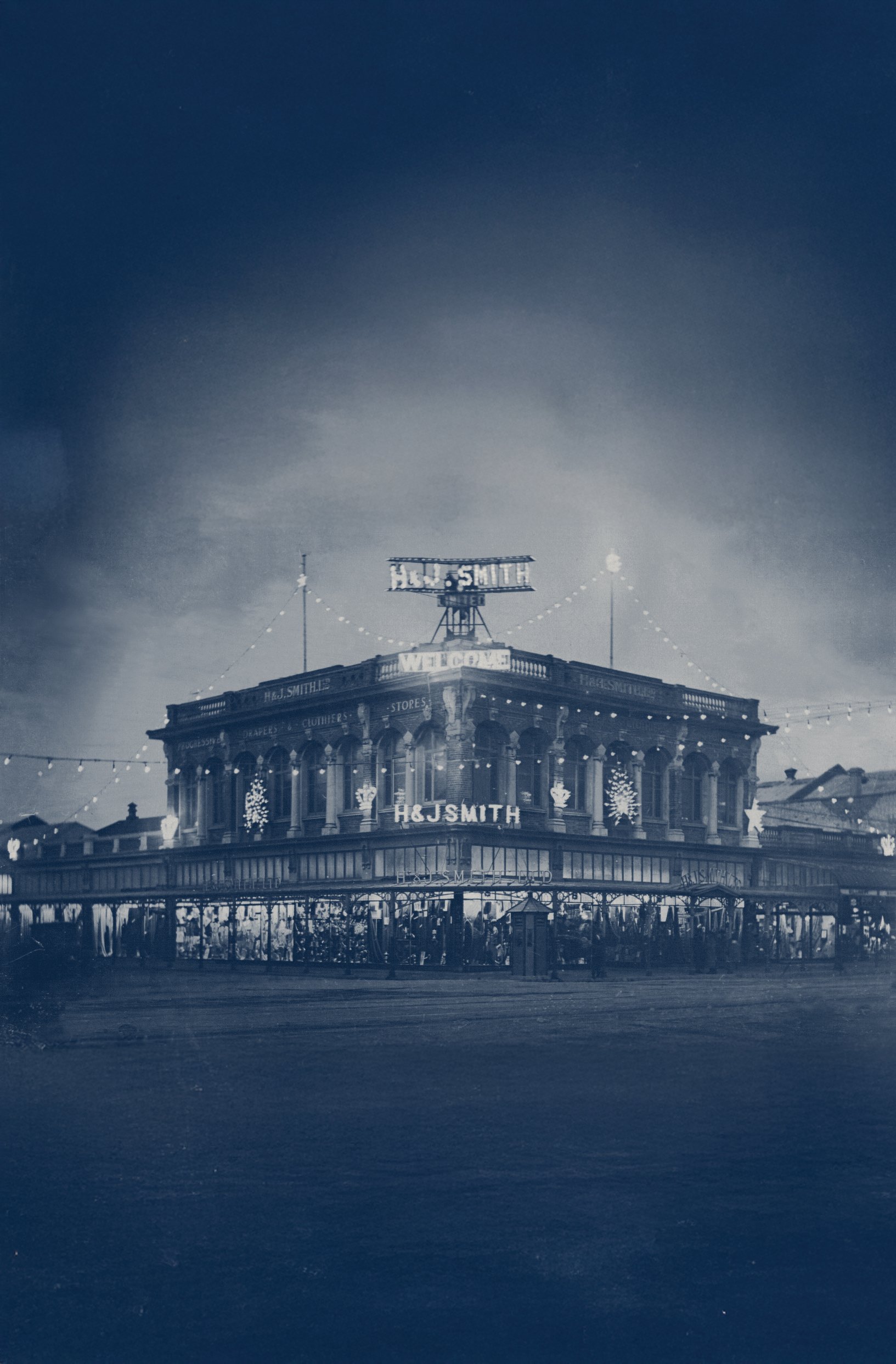 Black and white photograph of a vintage two-story building decorated with string lights and holiday ornaments, with signs reading "H&J Smith" and "Welcome."