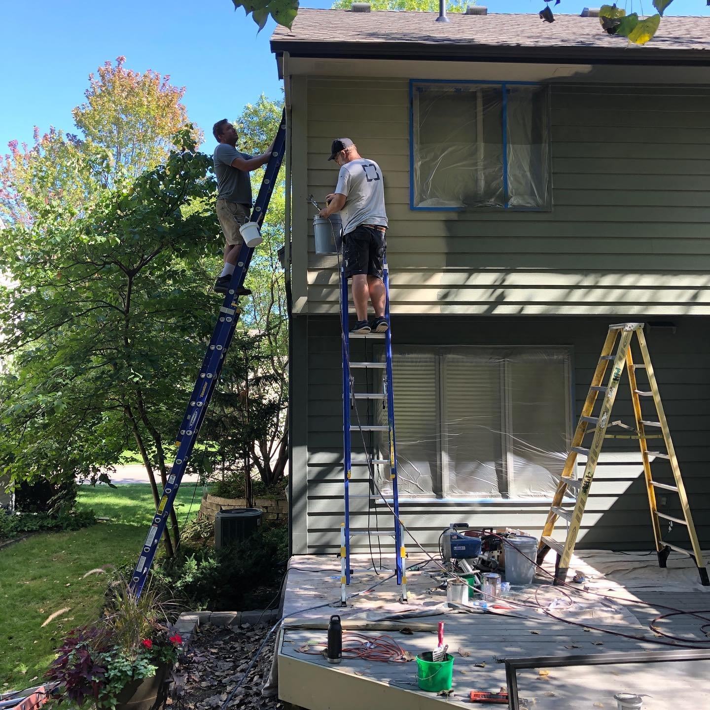 Two men working on the exterior siding of a two-story house, standing on ladders, with tools and paint supplies nearby, during daytime.