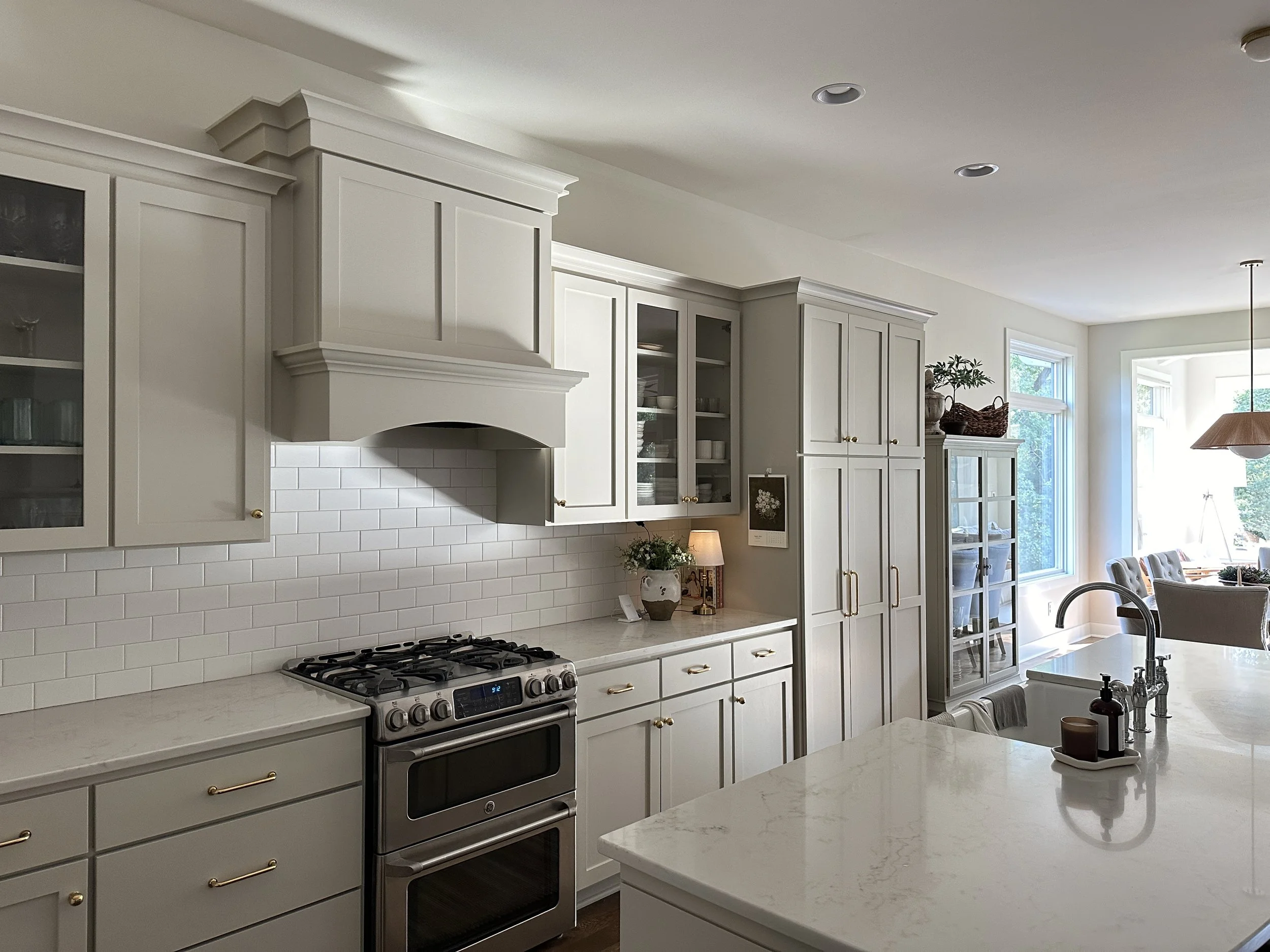 A bright, modern kitchen with white cabinets, a marble countertop, and a stainless steel stove. There are large windows letting in natural light, and a dining area with chairs is visible in the background.