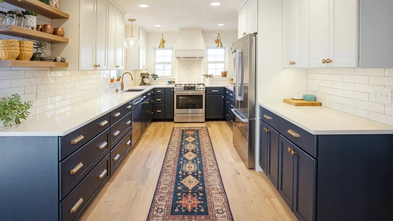 Kitchen with white upper cabinets and dark lower cabinets, white countertops, stainless steel appliances, a patterned runner rug, open wooden shelves with dishes and plants, and a white brick backsplash.