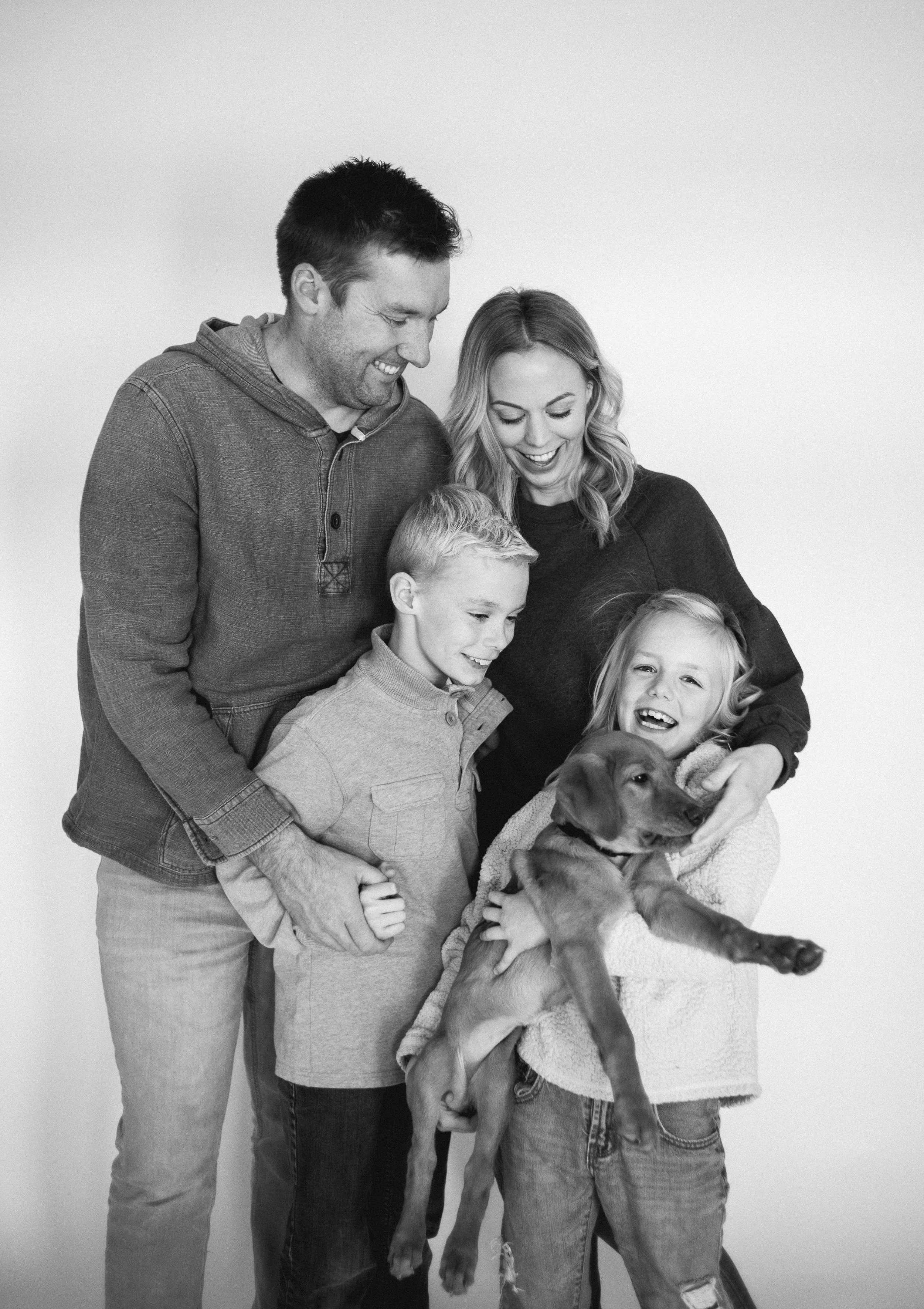 A family of five with two adults and three children, holding a puppy, smiling and looking at each other, against a plain background, in black and white.