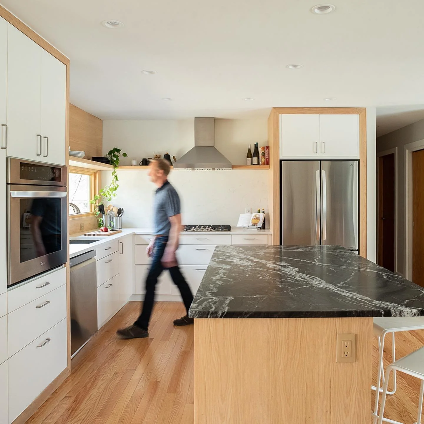 Modern kitchen with white cabinets, stainless steel appliances, black marble countertop, and wooden flooring. A person is walking through the space.