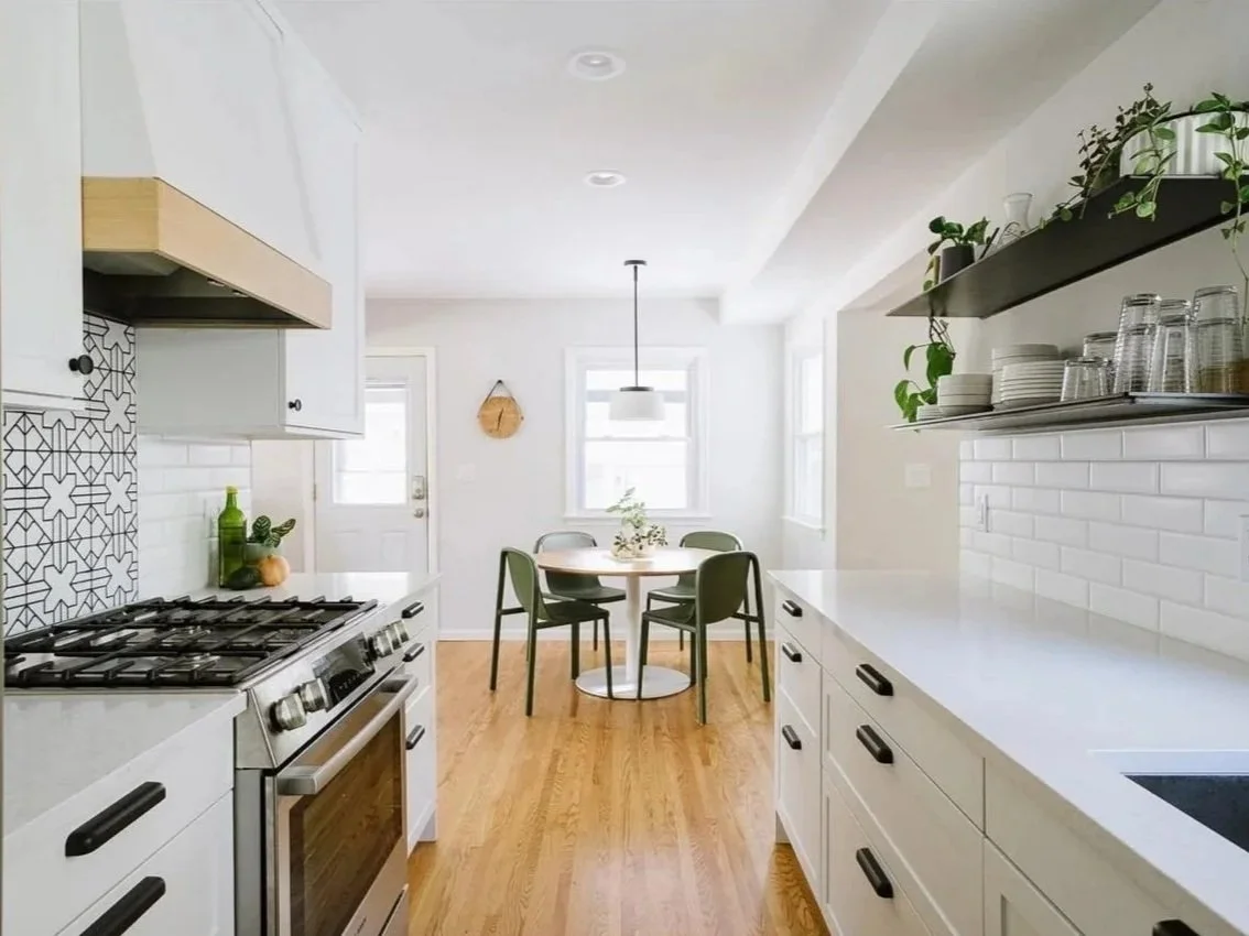 Bright, modern kitchen with white cabinets, wooden flooring, a round dining table with four green chairs, and decorative shelves with plants and dishes.