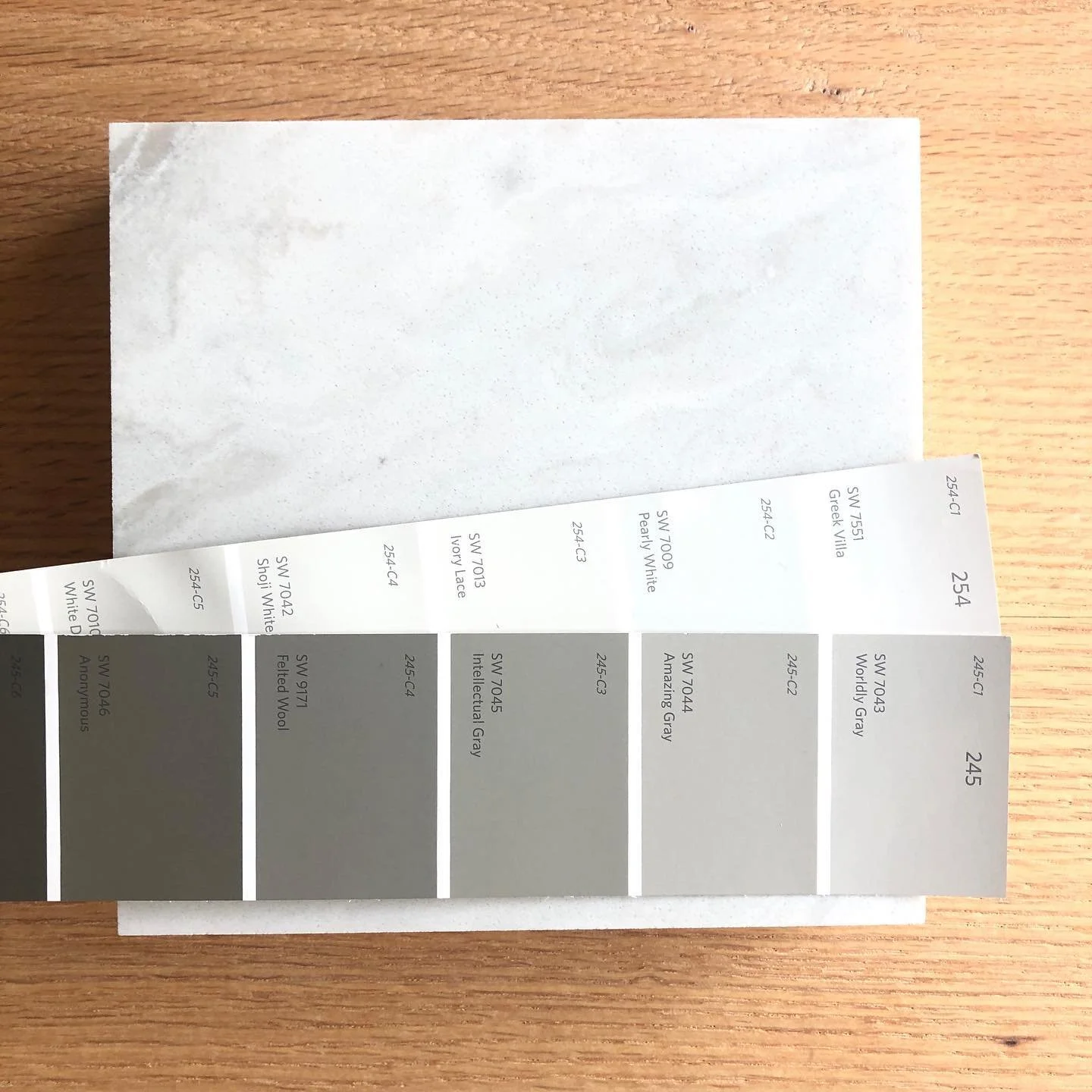 A color sample strip with shades of gray and white, placed on a wooden surface with an envelope and a paper behind it.