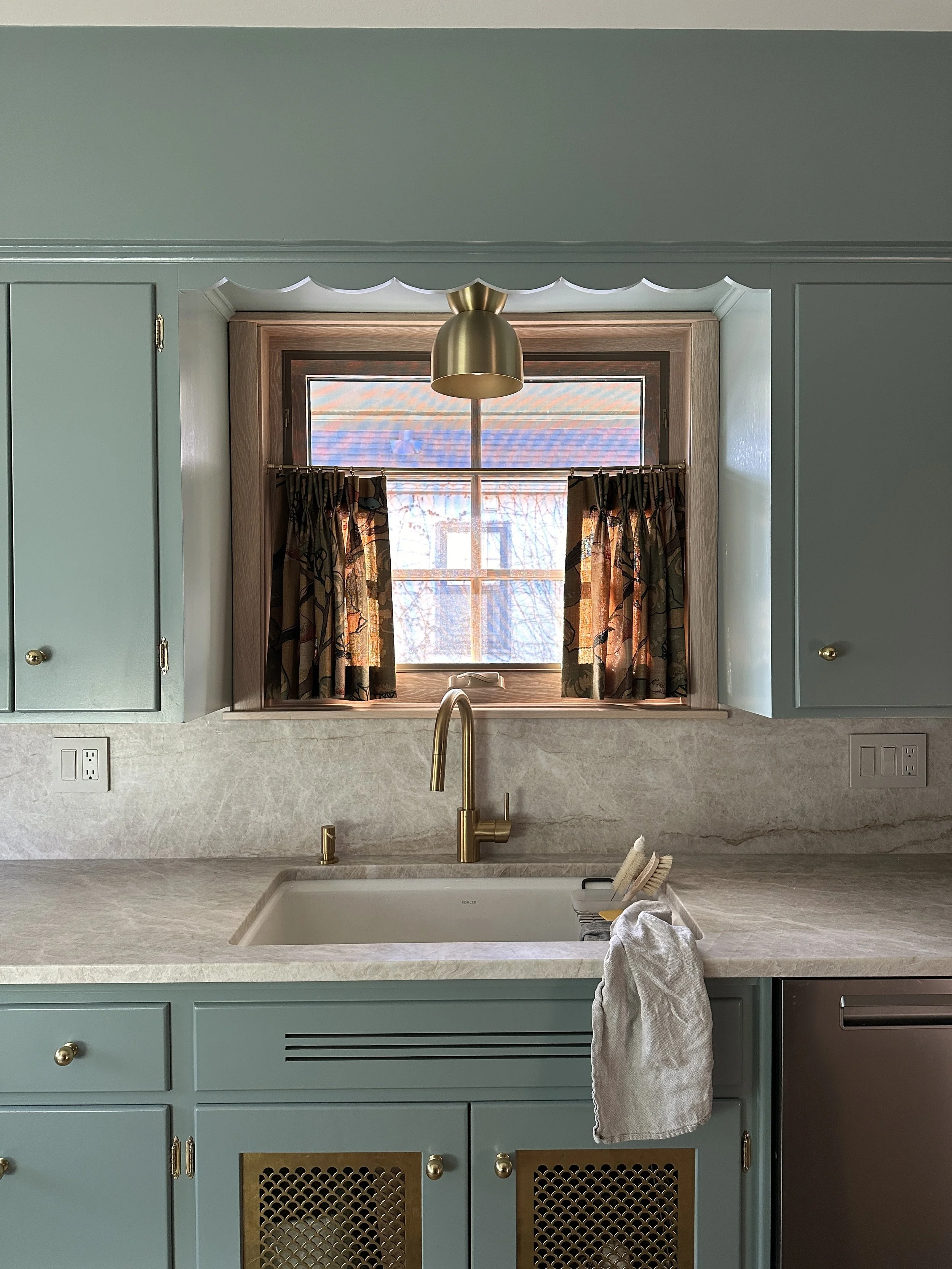 Kitchen with mint green cabinets, cream marble countertop, brass faucet, window with floral curtains, brass pendant light, and placed cleaning brushes.