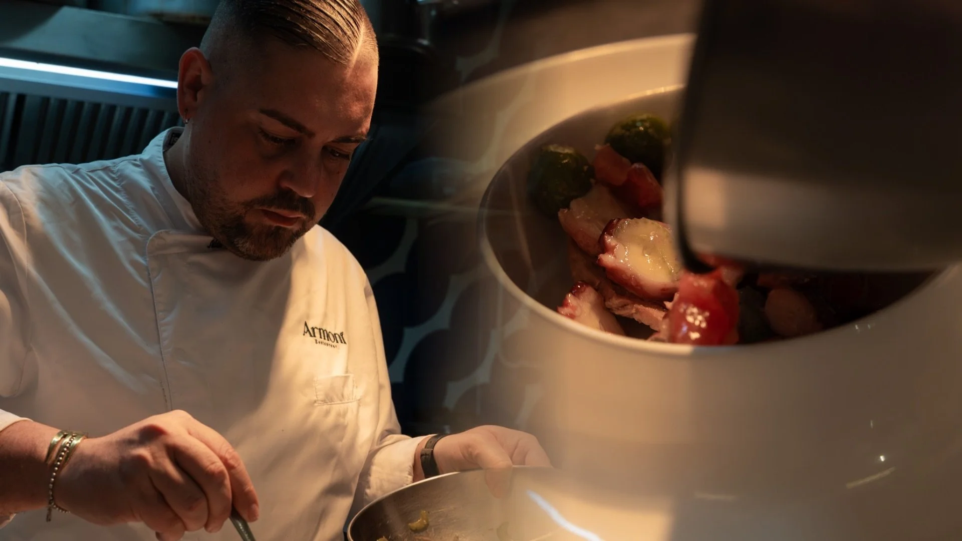 Uno chef in uniforme bianca controlla un piatto di cucina, mentre sullo sfondo si intravede una pentola con ingredienti