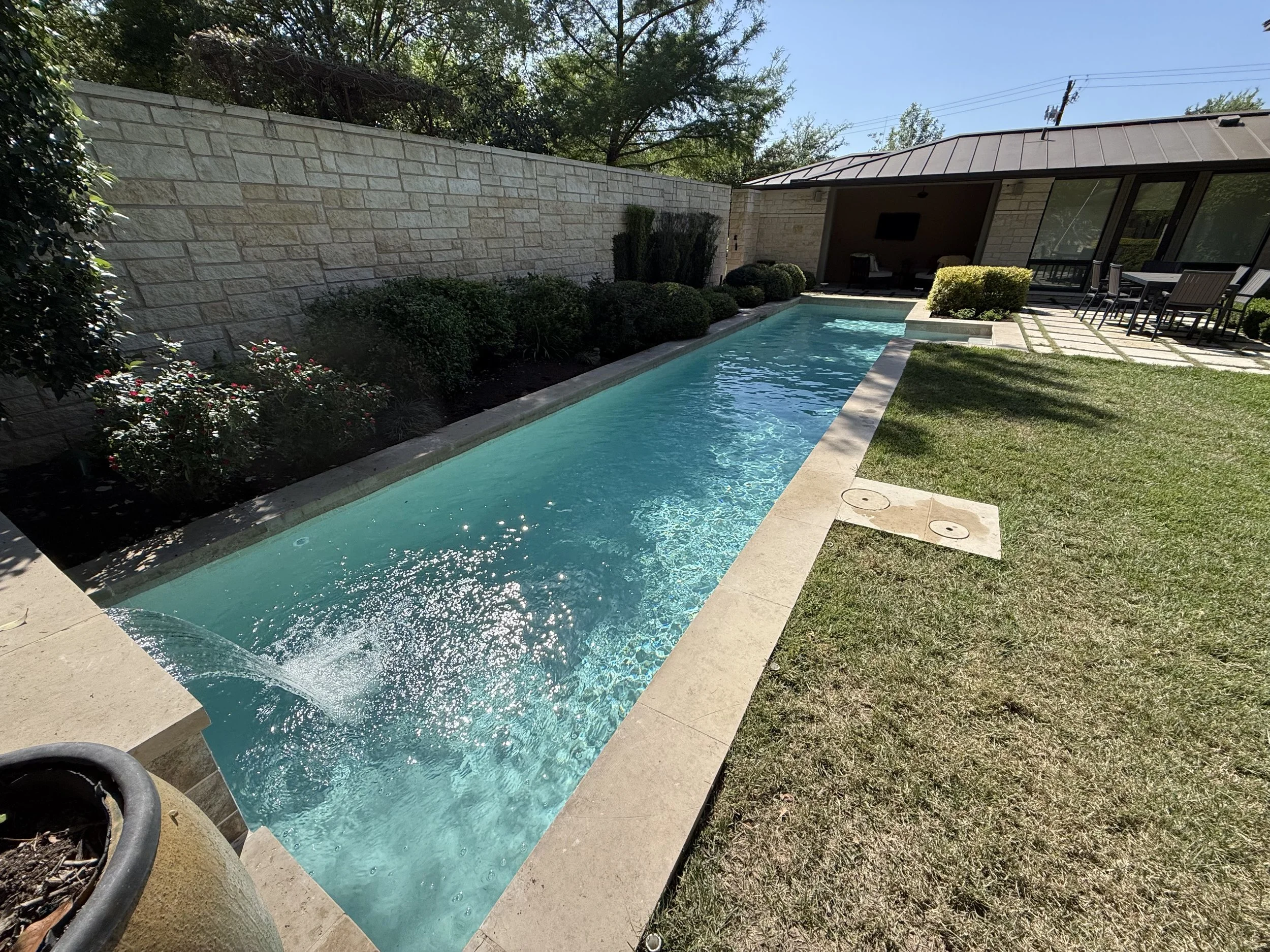 A narrow rectangular backyard swimming pool with clear blue water, surrounded by a stone deck, lush green bushes, and a grassy lawn, with a patio area featuring outdoor dining furniture and a covered outdoor living space in the background.