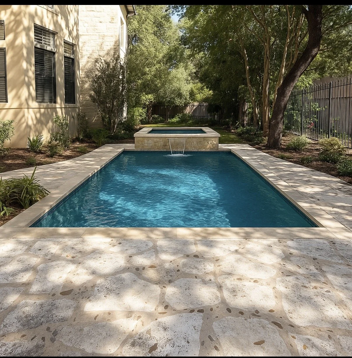 A backyard with a rectangular swimming pool and a raised hot tub, surrounded by a stone patio and lush trees and bushes, with a beige house wall and windows on the left and a black metal fence on the right.