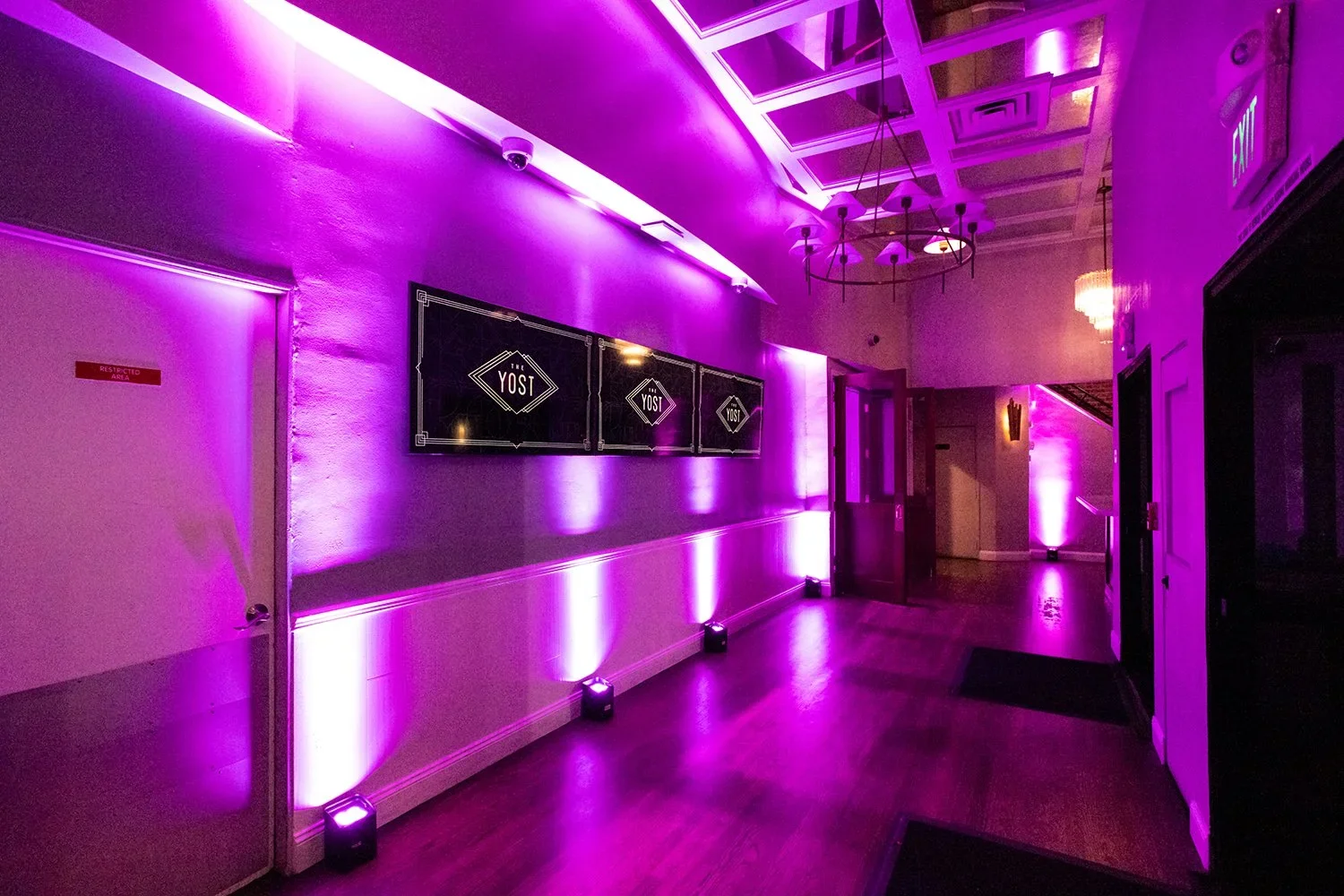 An indoor hallway illuminated with purple lighting, featuring three digital screens displaying the word 'YOST' mounted on a wall, and a chandelier hanging from the decorated ceiling.