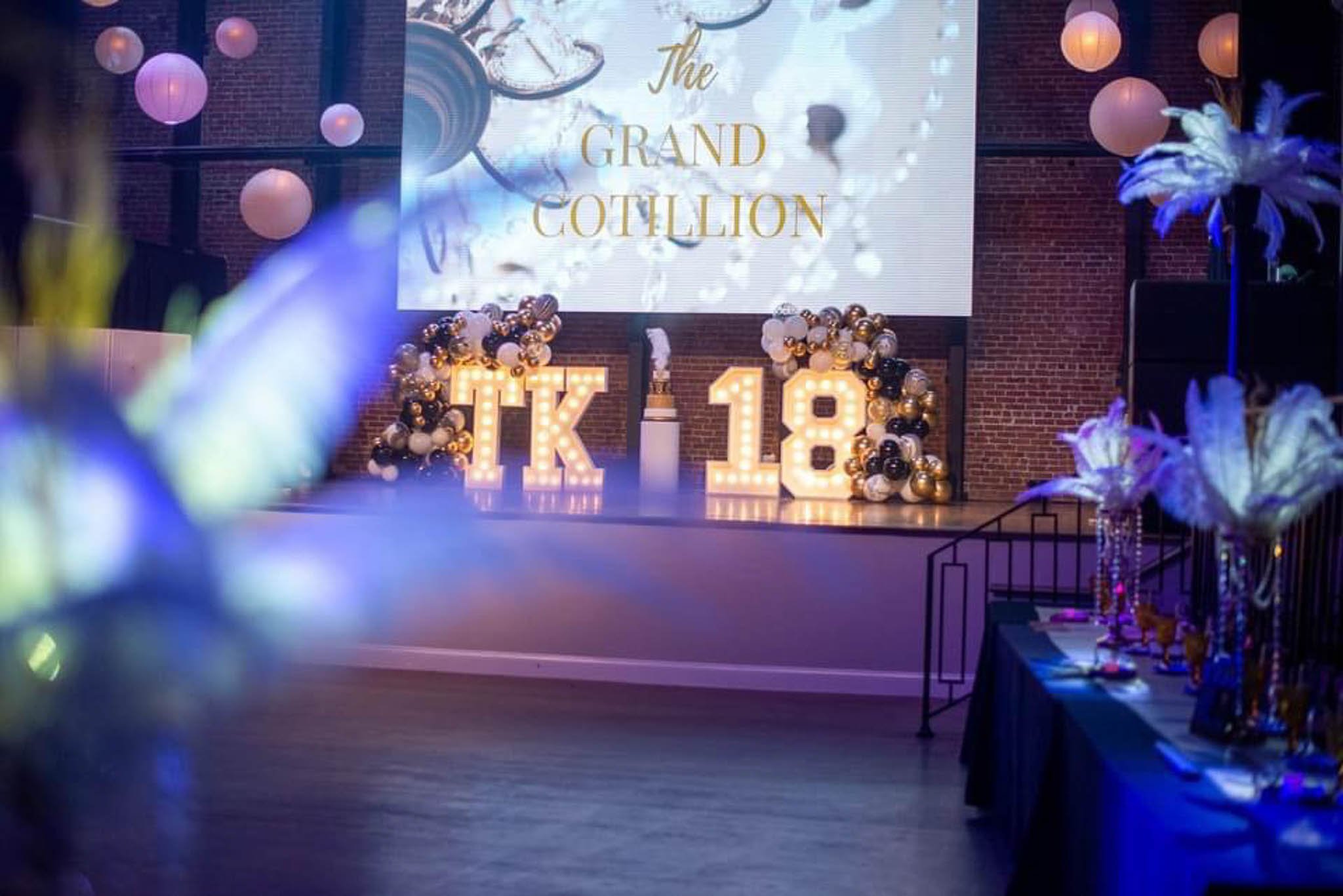Event stage with large illuminated decorations, balloons in black, white, and gold, a giant letter 'K' and number '18', and a large screen displaying 'The Grand Gouthlon'.
