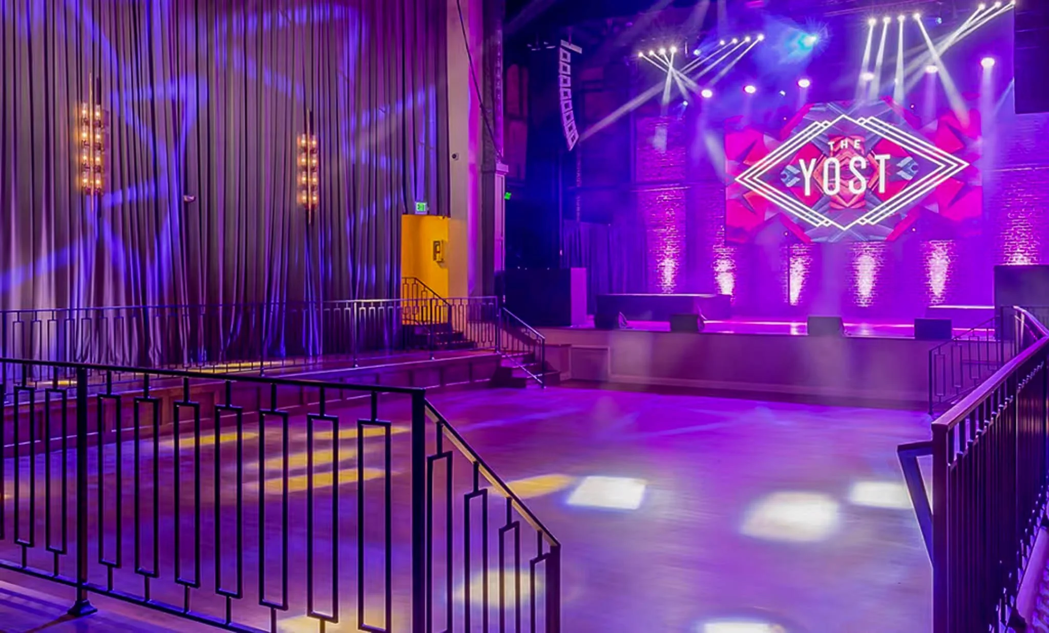 Empty stage with purple and pink lighting, large digital screen displaying 'THE YOST', surrounded by brick walls and purple curtains, in an auditorium or event space.