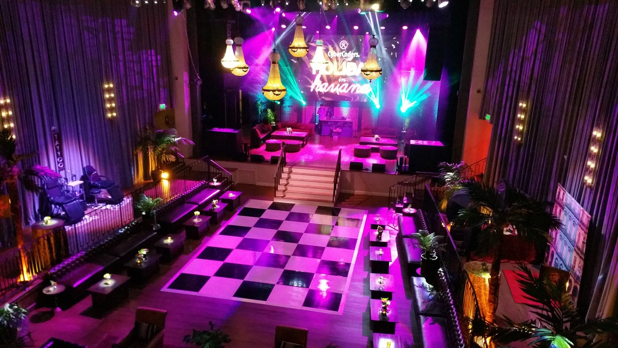 An indoor event space with a stage illuminated by colorful lights, chandeliers hanging from the ceiling, and black-and-white checkered flooring. Small tables with flowers are arranged around the dance floor.