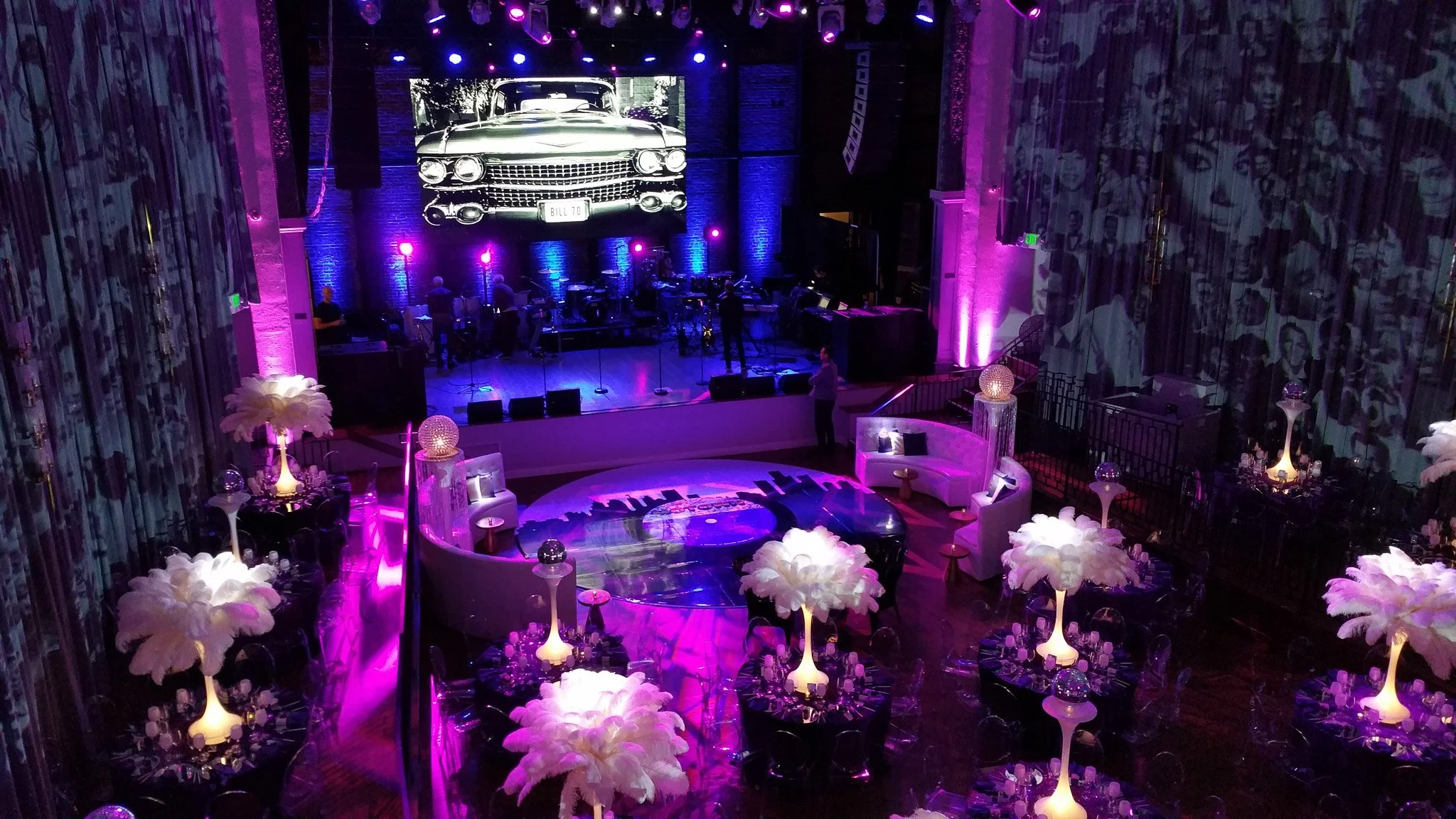 A stage with musical instruments set up, a large screen displaying a vintage car with a license plate reading 'BILL 70', and pink and purple lighting. In the foreground, there are white fluffy flowers on small tables and a circular sofa surrounding a graphic on the floor.