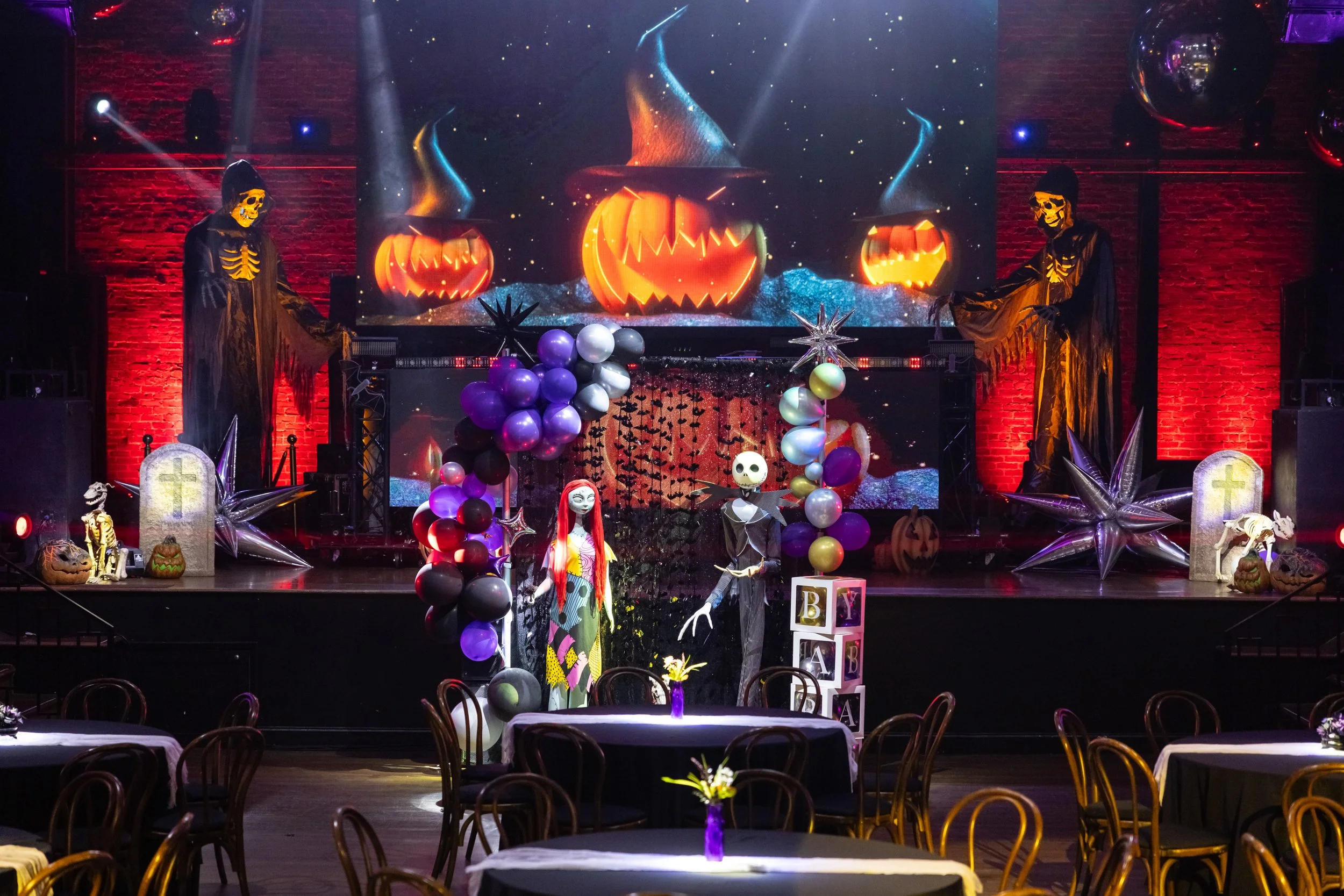 Interior of a venue decorated for Halloween with balloons, skeletons, pumpkins, and a large digital screen showing glowing jack-o'-lanterns and witch hats.