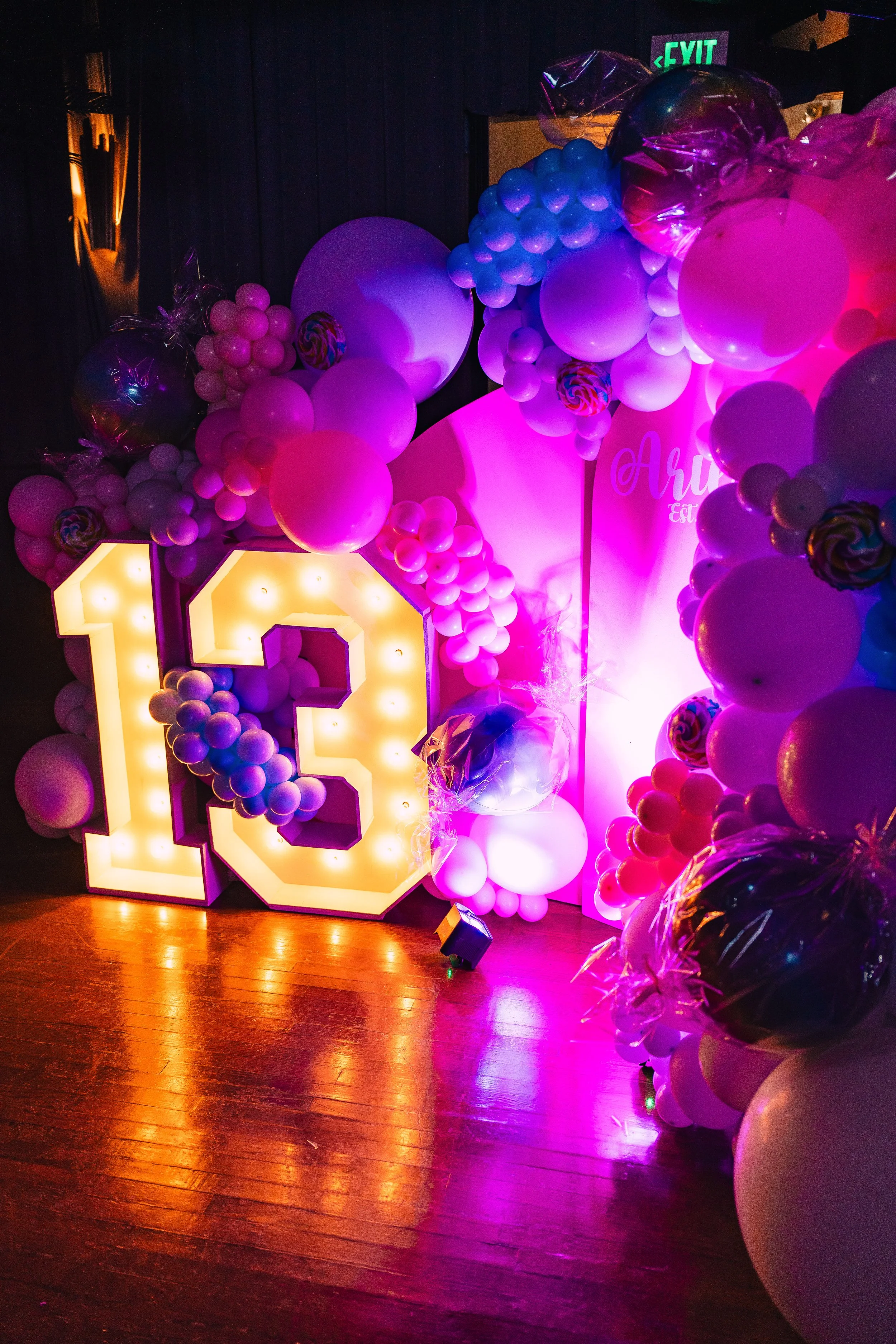 A decorated birthday celebration area with a large illuminated sign showing the number 10, surrounded by pink, purple, and white balloons, some wrapped in clear plastic, and a pastel pink backdrop with cursive writing.