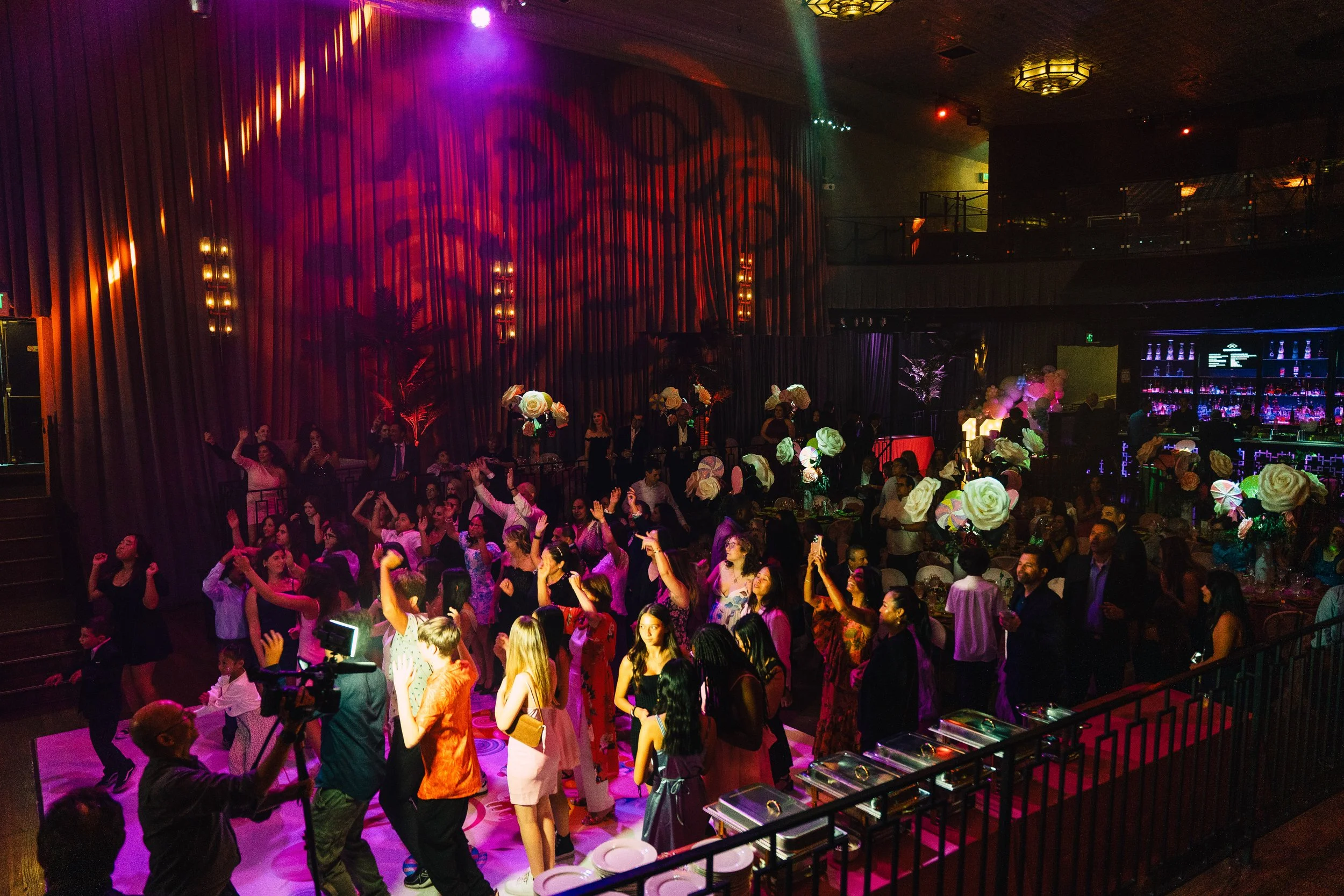 A crowded indoor party scene with colorful lighting. People are dancing on a dance floor, many wearing festive clothing and hats. The room is decorated with large white flowers and balloons. The background features a bar area with illuminated bottles and drink glasses.