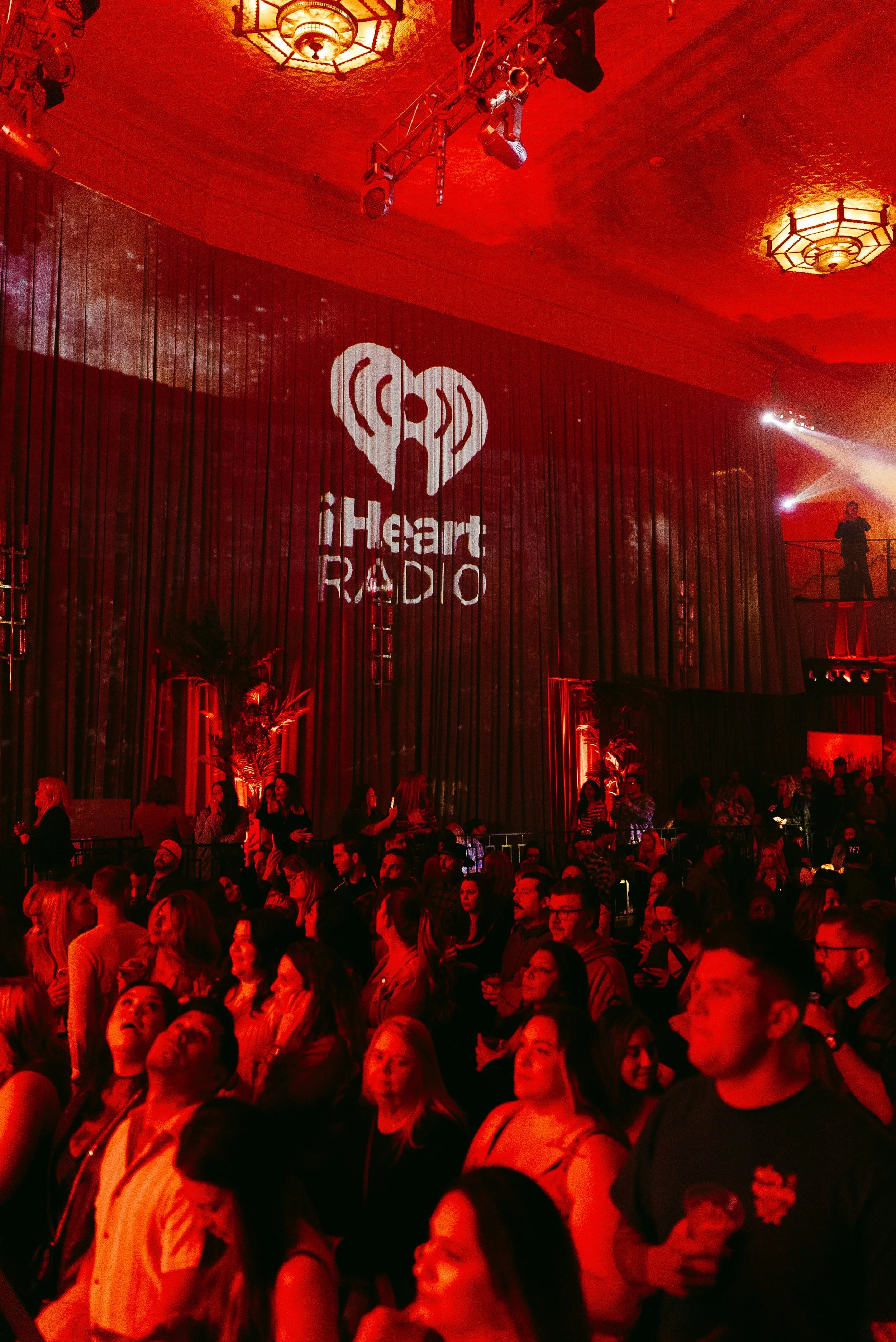 Crowd of people at a concert or event, illuminated with red lighting, with a large screen displaying the iHeartRadio logo.