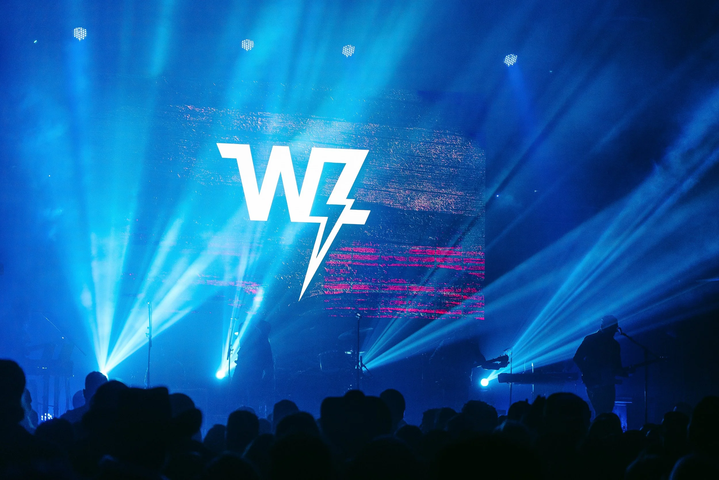 A concert stage illuminated with blue lights and a large screen displaying the 'WZ' logo with a lightning bolt, with silhouettes of people and equipment in the foreground.