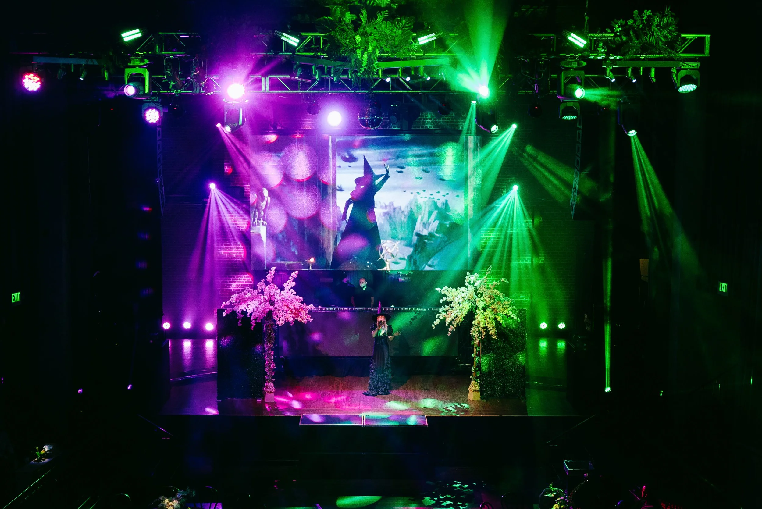 A stage with colorful pink, purple, green, and blue lighting, featuring dancers in silhouette and floral decorations, with a musician playing a keyboard at the front.