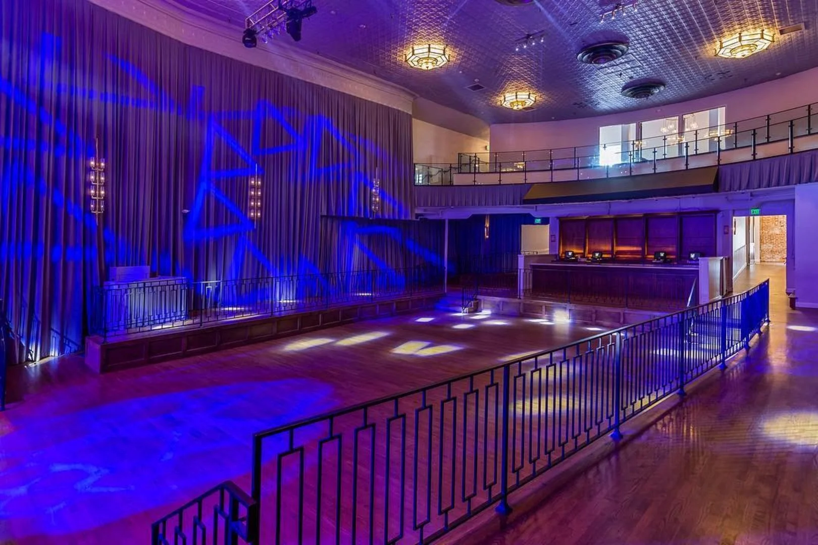 Empty indoor event space with a wooden dance floor, decorated with blue lighting and star-shaped projections, surrounded by a railing, with a stage and reception desk visible.