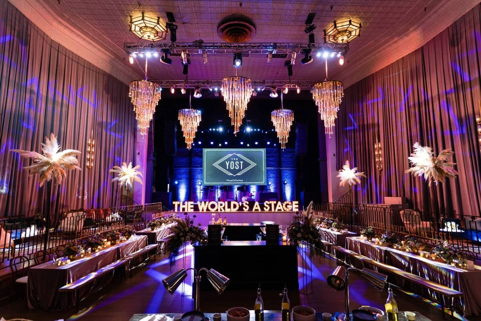 An event venue decorated for a performance or celebration, with a stage, large sign saying 'The World's a Stage,' elegant chandeliers, palm tree decorations, tables with floral centerpieces, and a large screen displaying 'The YOST' logo.