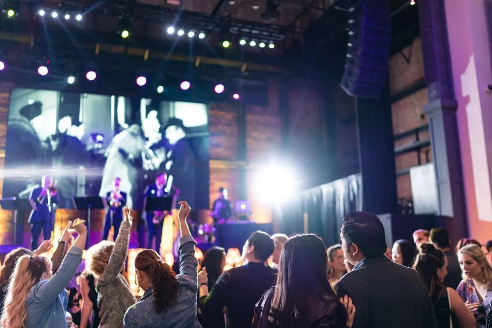 A crowd of people at a concert or event, facing a stage with bright lights and a large black-and-white screen showing figures of a performer and musicians.