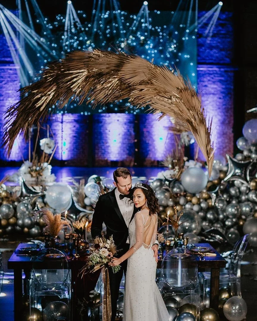 A bride and groom pose together at their wedding reception decorated with balloons and festive lighting.