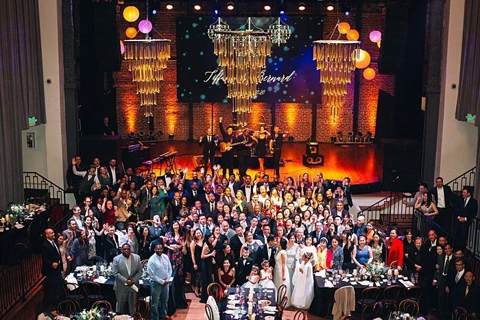 Large wedding celebration with many guests, a musical band on stage, elegant chandeliers, colorful hanging paper lanterns, and decorated tables.