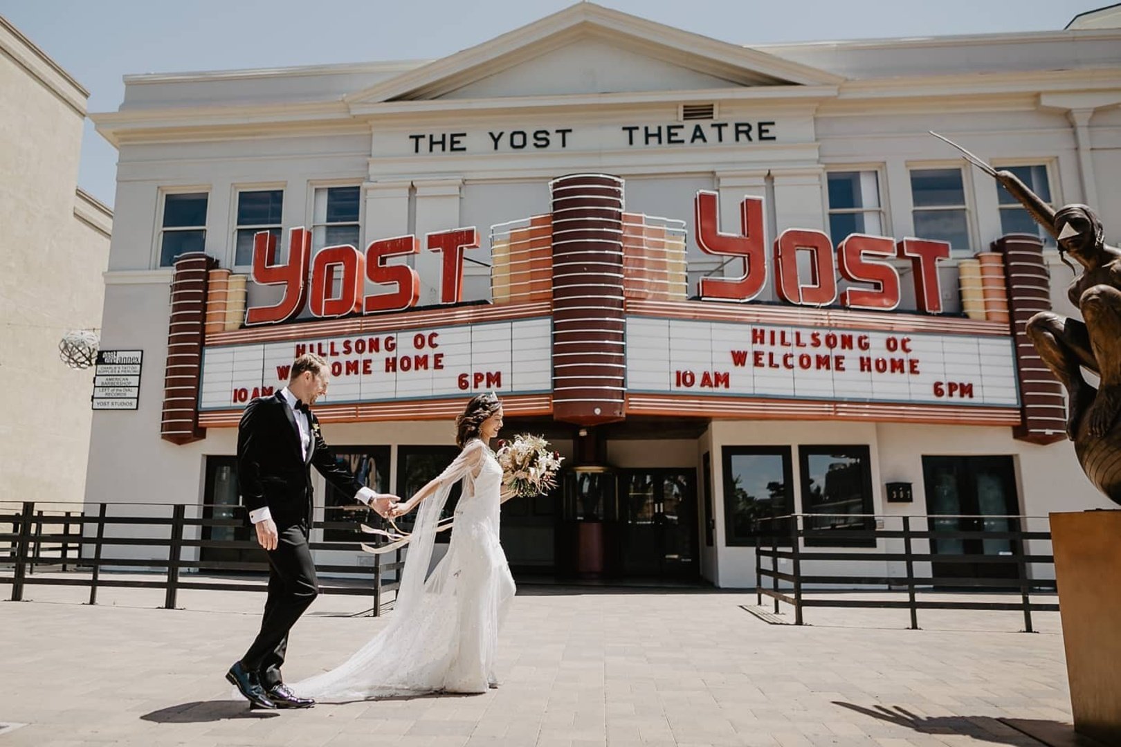 A bride and groom holding hands outside the Yost Theatre, which has a marquee displaying event times and titles.
