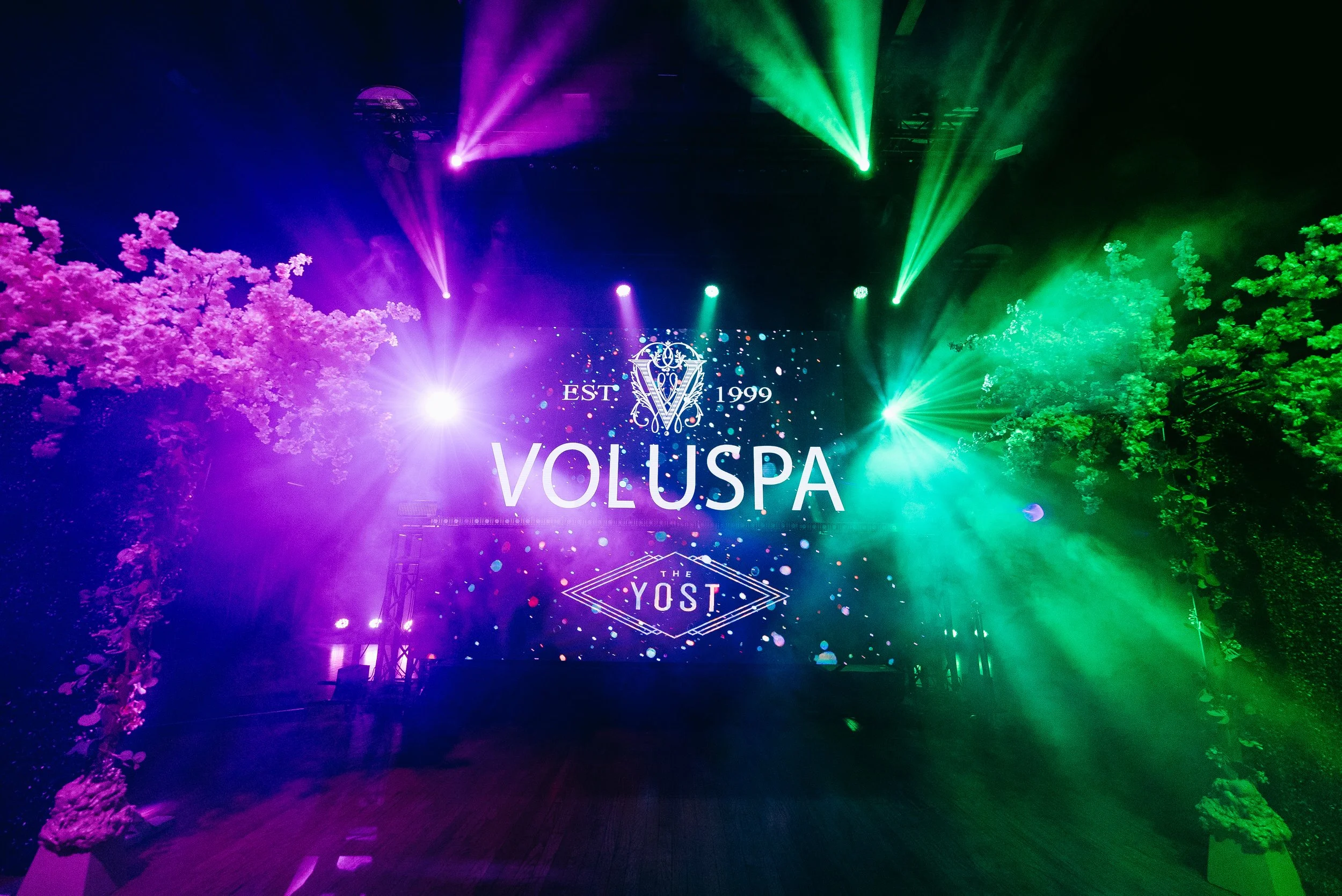 Colorful nightclub stage with pink and green lighting, decorative trees, and a large screen displaying 'VOLUSPA' and 'YOST' logo.