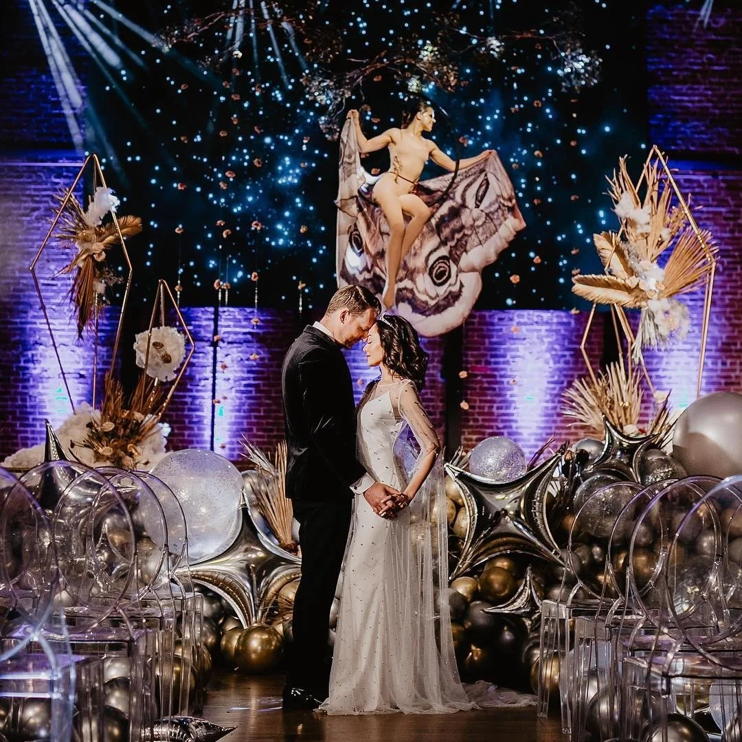 A couple in formal attire standing close, holding hands, with a decorated wedding or event backdrop featuring balloons, dried floral arrangements, and artistic lighting. There is a large butterfly and a performer in a fantasy-themed costume leaping i