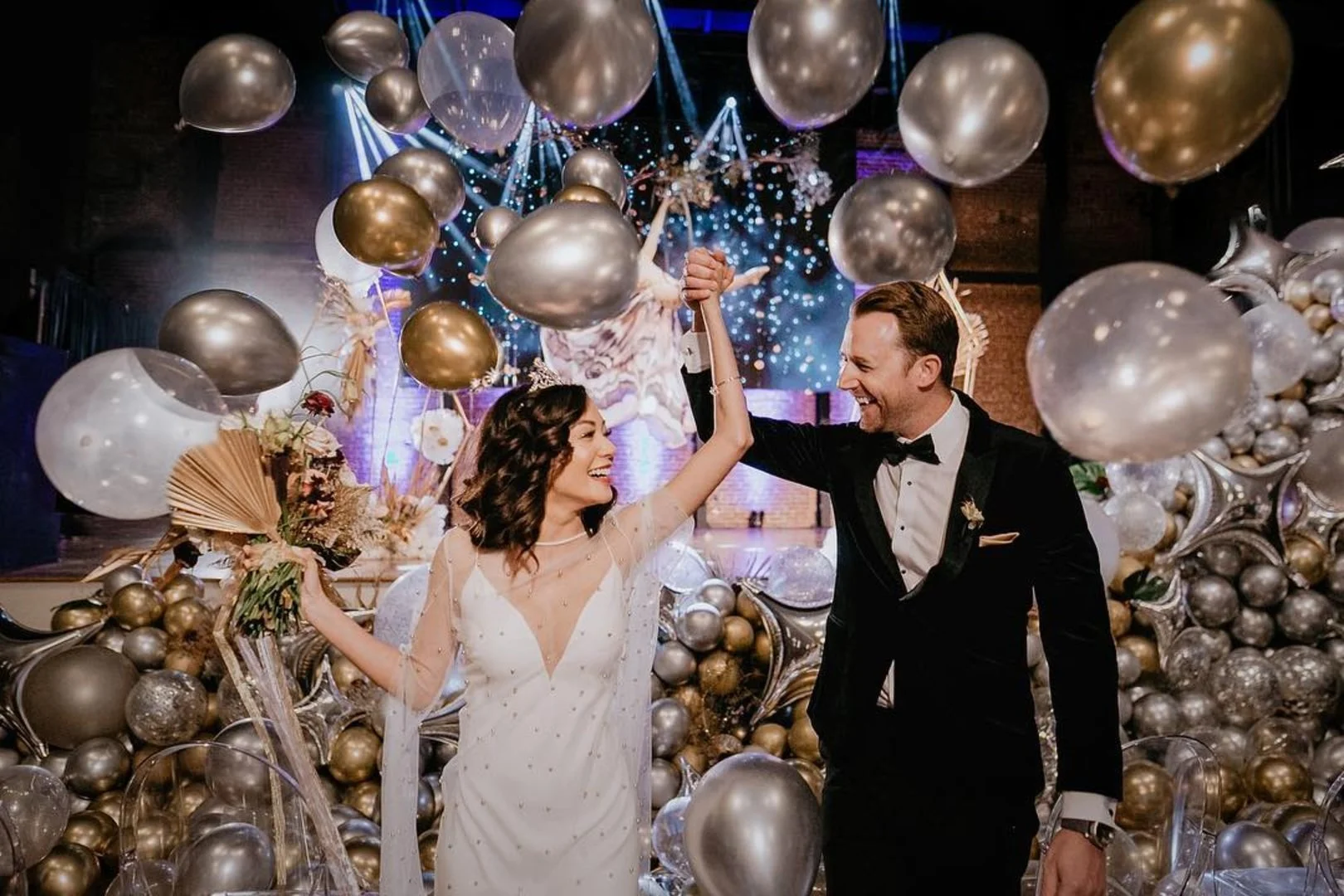 A bride and groom dancing at their wedding reception surrounded by silver, gold, and clear balloons and decorations, with a starry backdrop and festive lighting.