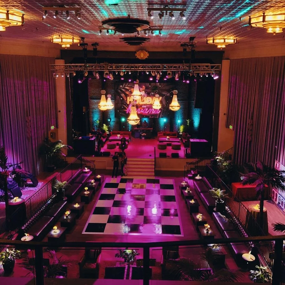Elegant event space with a dance floor, tables with flowers and candles, and a stage with lighting and a neon sign that says 'Flora in Miami.'