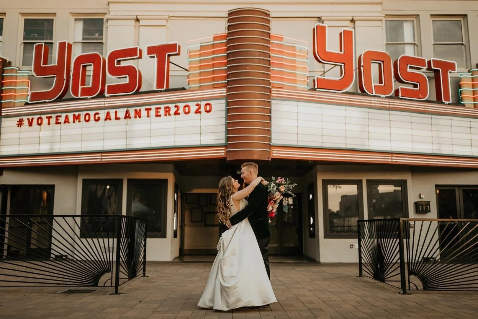A couple in wedding attire hugging in front of a vintage movie theater with the words 'YOST' on the marquee and '#VOTEAMOGALANTER2020' written on it.