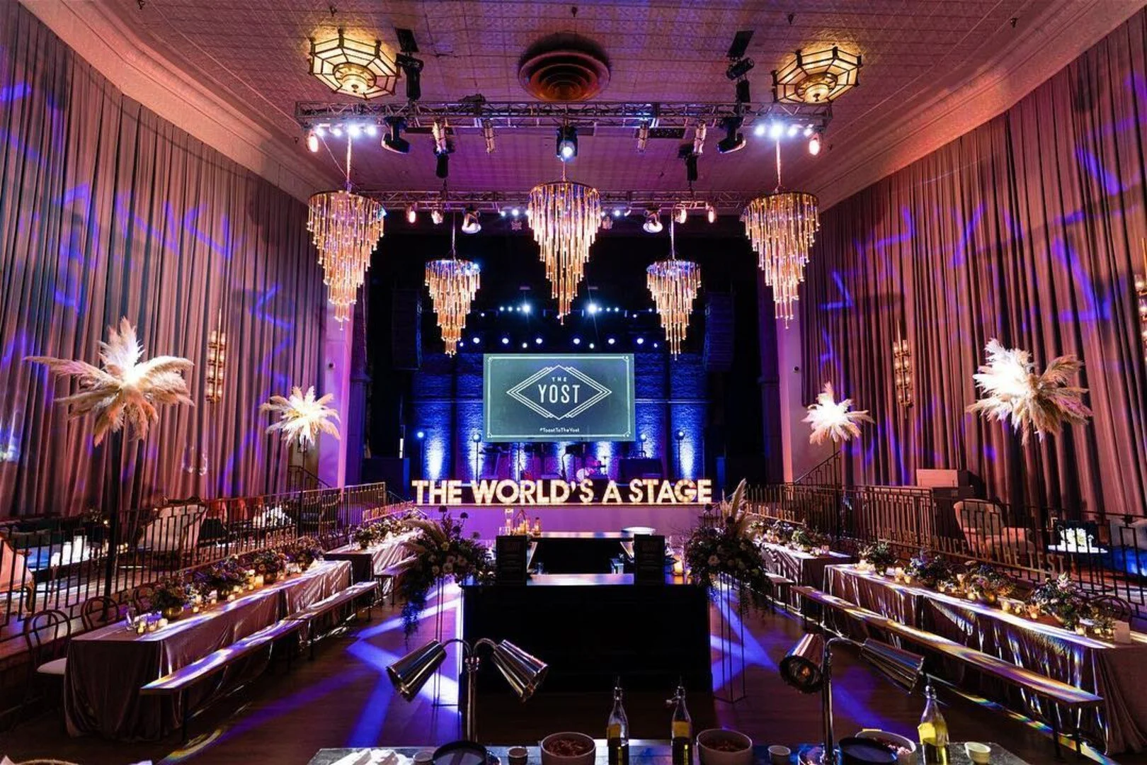 A decorated event stage with large chandeliers, palm tree decorations, and hanging lights. There are long tables with flower arrangements, and a sign that reads 'THE WORLD'S A STAGE' in front of the stage. The stage has a big screen displaying 'YOST'