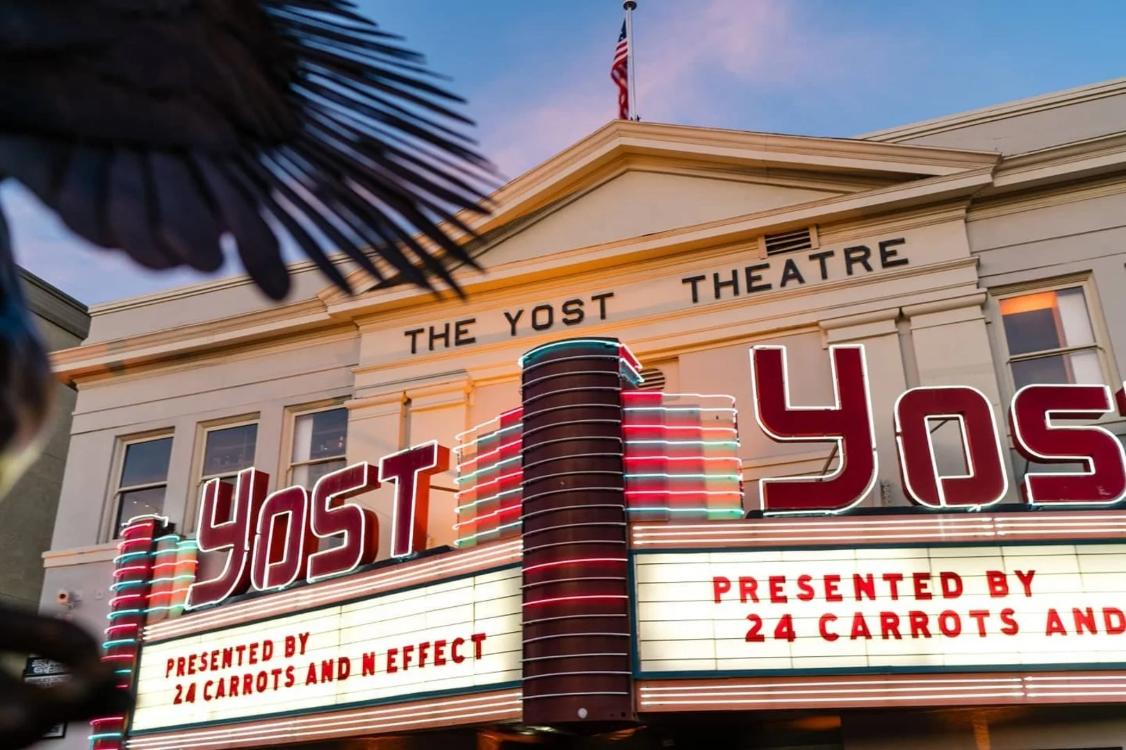 The marquee of the Yost Theatre with neon lights spelling out YOST, the building's sign reads "The Yost Theatre" and there is an American flag on top. The marquee also displays text about a show presented by 24 carrots and N Effect.
