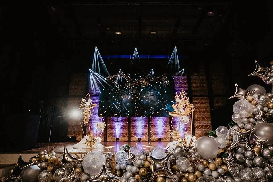 Stage decorated with gold and silver balloons, large ornamental plants, and a digital screen showing a galaxy scene with spotlights shining down.