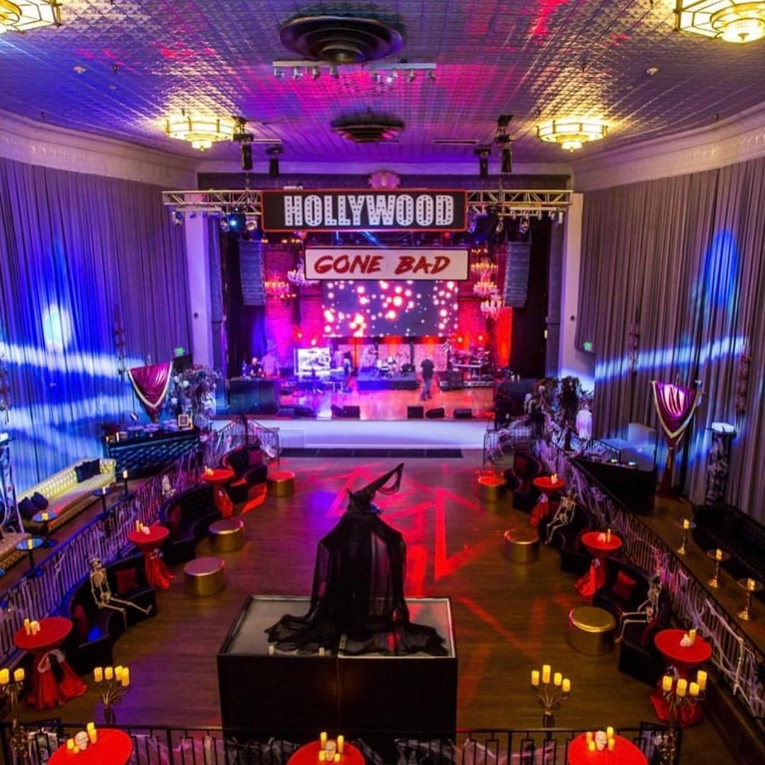 Decorated event venue with a stage displaying a screen and signs reading 'HOLLYWOOD GONE BAD.' The room has tables with red tablecloths, candles, and skeleton decorations, and a black mannequin dressed in dark clothing and veil is displayed in the ce