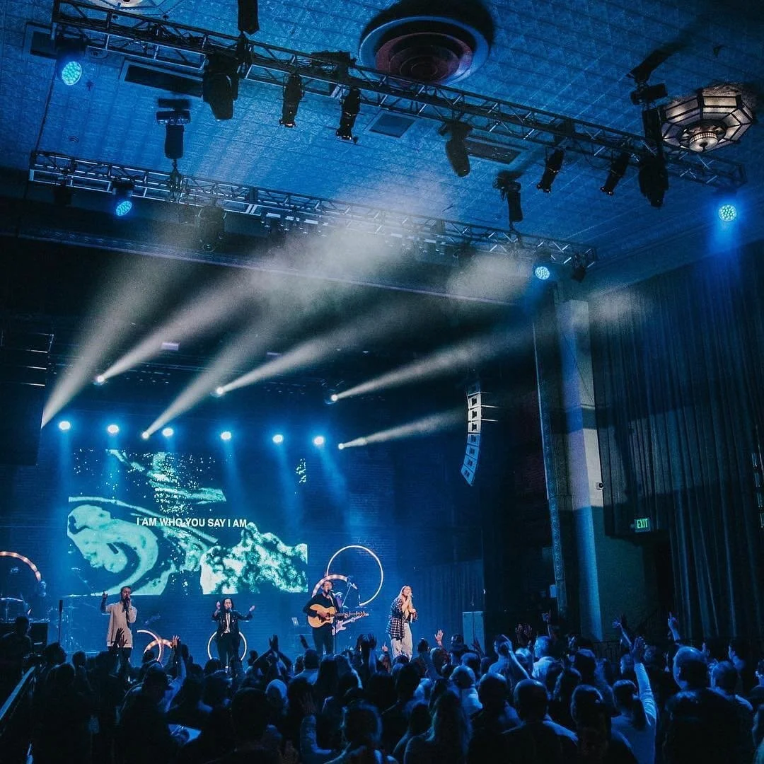A live concert with a band performing on stage, audience raising their hands, illuminated by blue stage lights, and a digital backdrop showing water and the text 'I AM WHO YOU SAY I AM'.
