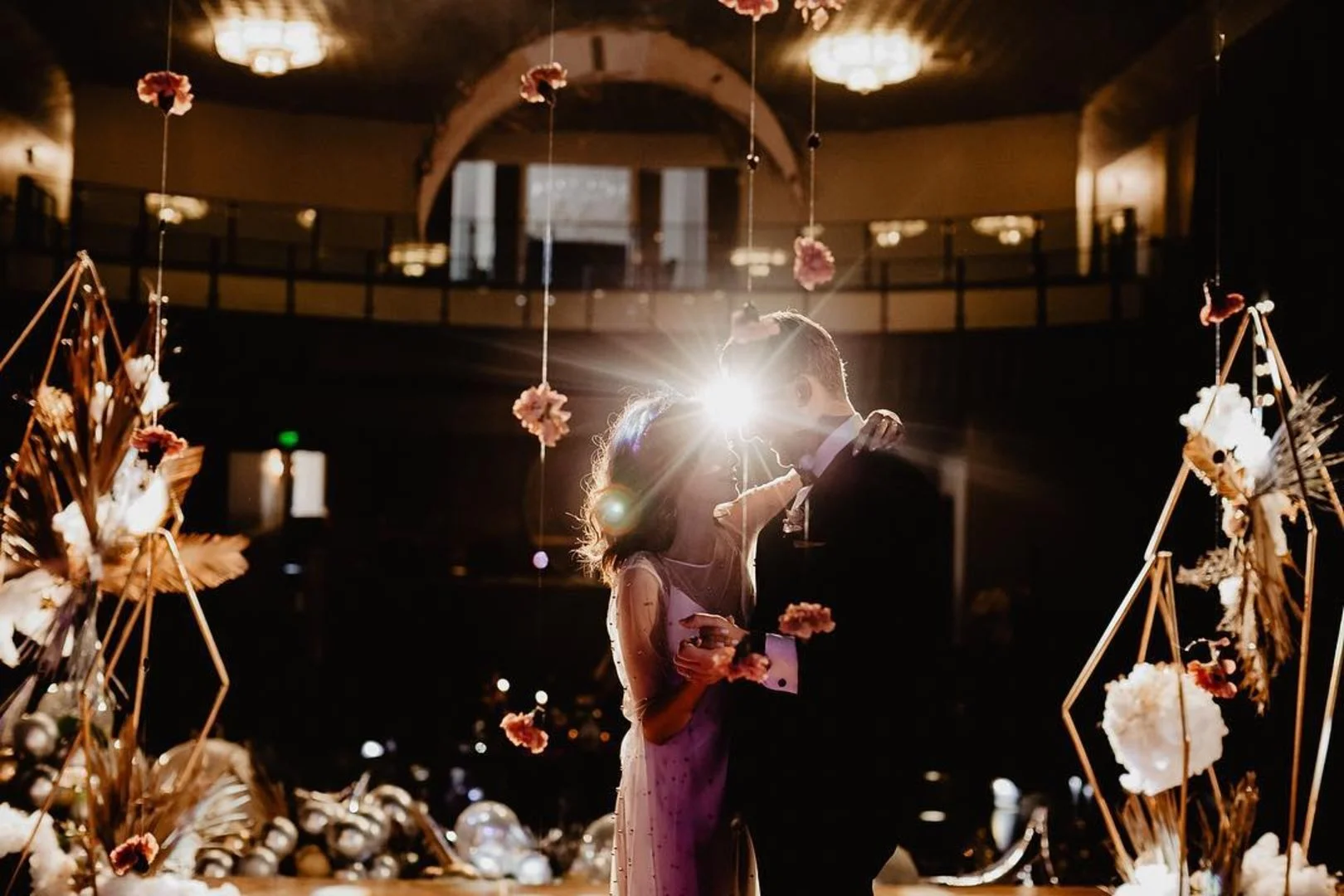 A couple dances closely during their wedding reception, with bright sunlight shining behind them. They are surrounded by floral decorations and elegant lighting in a decorated venue.