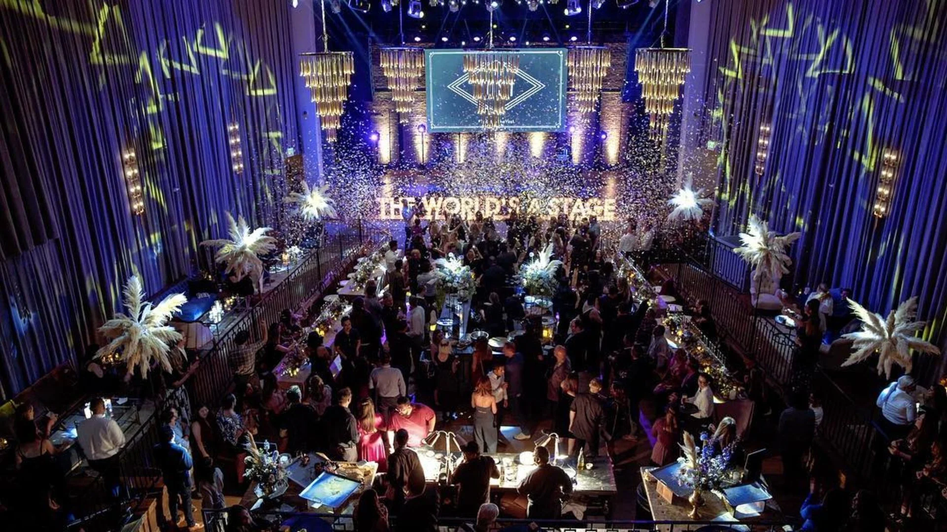 People dancing and socializing at a lively event with purple lighting, confetti falling, and elaborate decorations including large feather arrangements, chandeliers, and a sign that reads 'THE WORD POP UP STAGE' in the background.